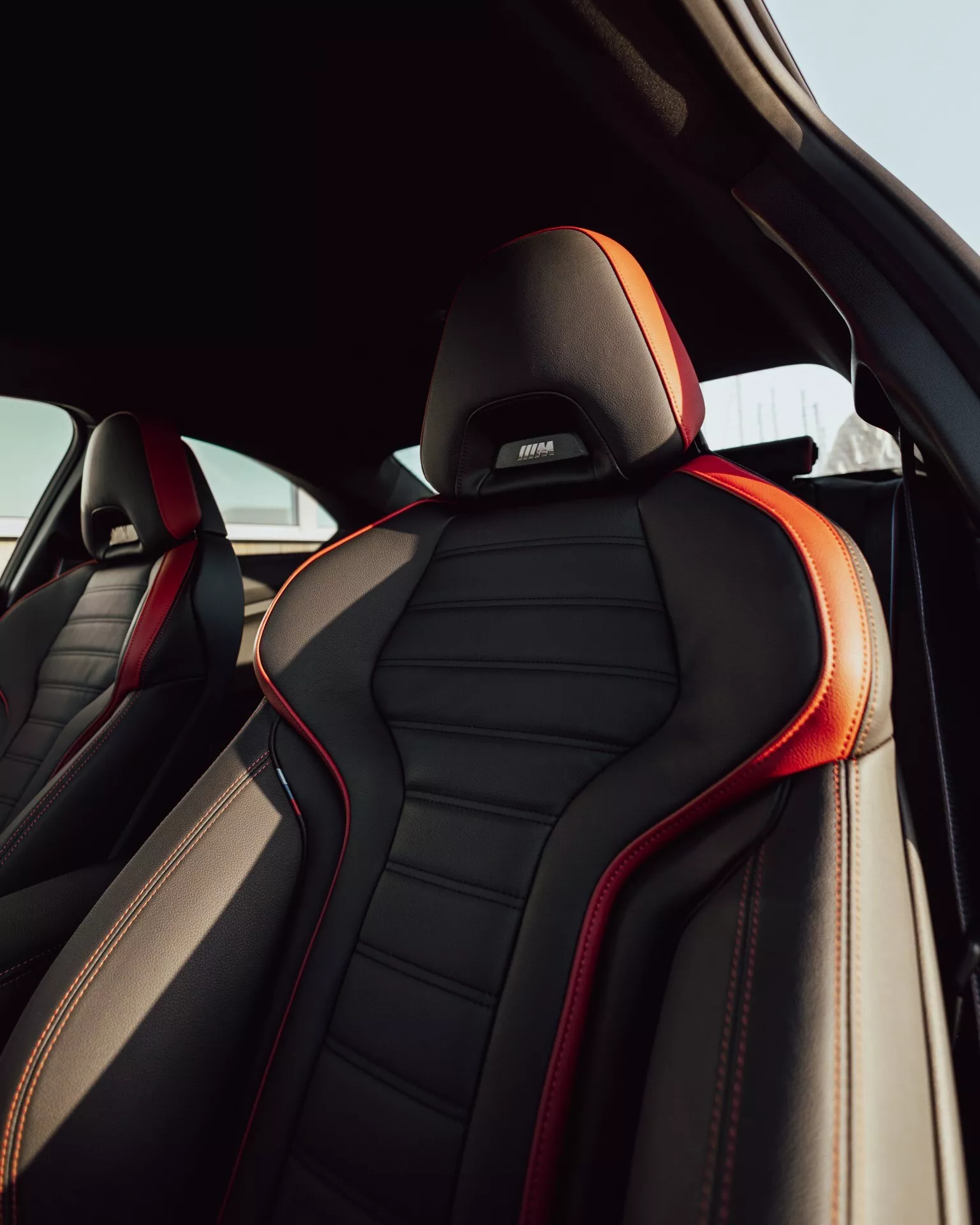 Detailný pohľad na predné sedadlo cestujúceho BMW 2er-Reihe 2024 (G42-G87 Facelift) zo zadného priestoru pre cestujúcich, zobrazujúci luxusné čierne kožené čalúnenie s červeným šitím a lemovaním. Ergonomický dizajn s výraznými bočnými výztuhami a opierkou hlavy s vyšívaným logom výkonnostnej alebo luxusnej podznačky, všetko v prirodzenom dennom svetle.