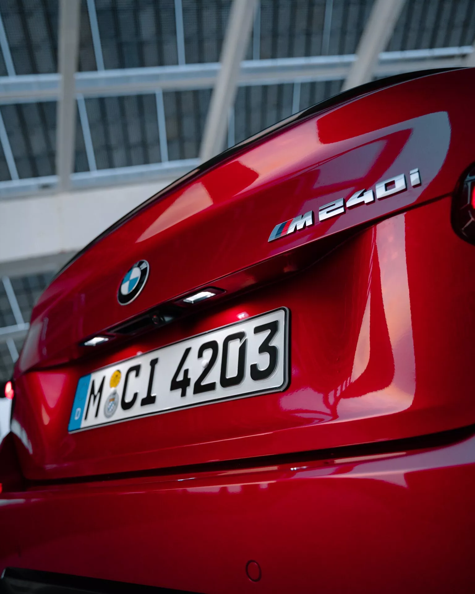 Detailný pohľad na zadnú časť červeného BMW M240i, zobrazujúci emblém BMW, označenie modelu, výklopné veko kufra, zadné svetlá v tvare L a integrovaný zadný spojler s mestským alebo priemyselným pozadím.