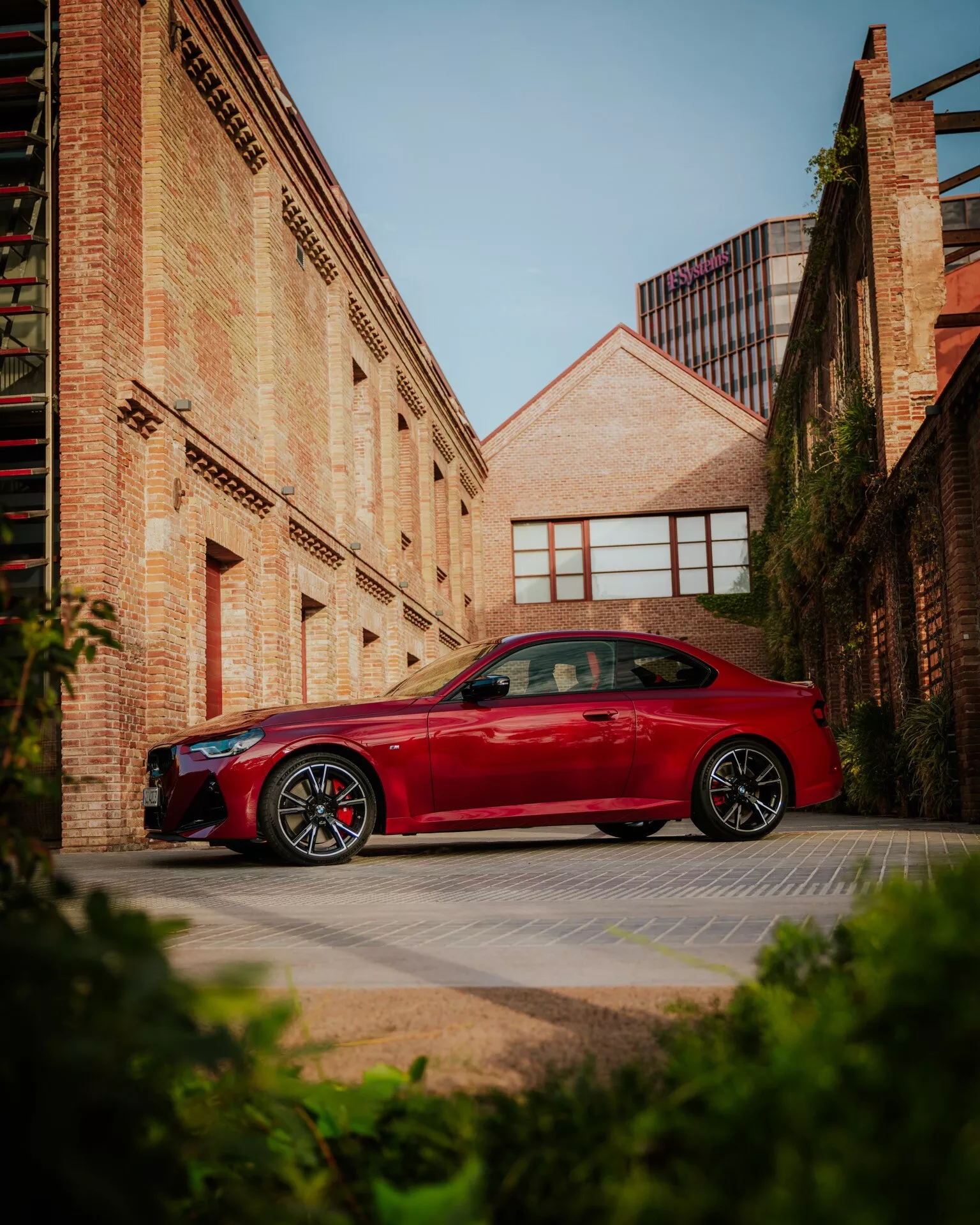 Červené BMW 2 série 2024 (facelift G42-G87) zaparkované v mestskom prostredí s tehlovými budovami v pozadí. Auto je zobrazené z profilu, s dizajnovými líniami cez dvere a blatníky a jasne viditeľnými športovými zliatinovými kolesami. Obrázok je vyhotovený z nízkeho uhla, čo zvyšuje impozantný postoj vozidla proti rannému alebo popoludňajšiemu slnku, ktoré vrhá jemné tiene. Táto fotka zachytáva témy automobilového dizajnu, športových úprav a mestského životného štýlu.