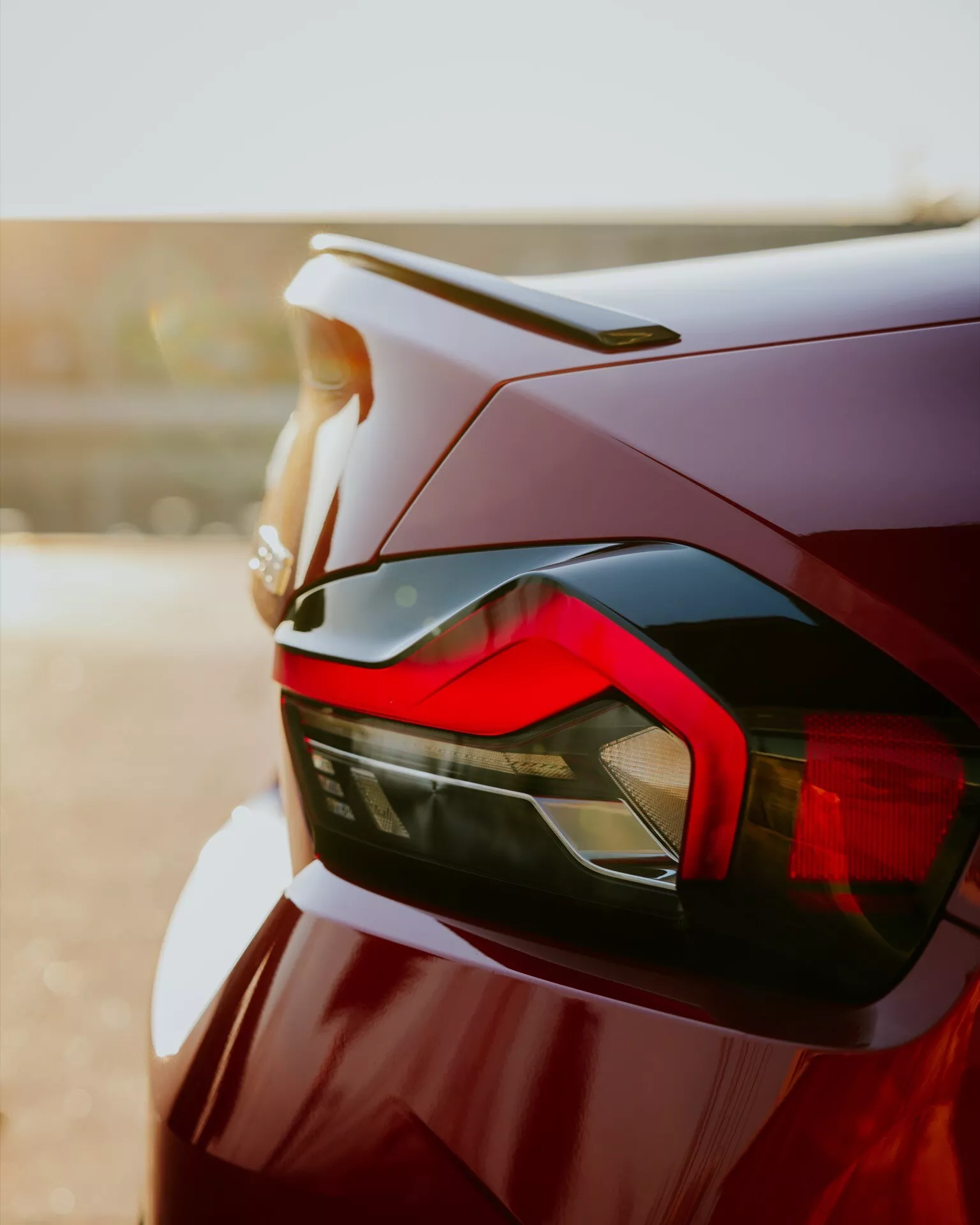 Detailný záber zadnej časti vozidla BMW 2er-reihe 2024 (G42-G87 facelift), zdôrazňujúci dizajnové prvky vozidla, ako je elegantné zadné svetlo s hranatými LED prvkami, jemný zadný spojler integrovaný do veka kufra a vysokokvalitný lak v tmavočervenej alebo vínovej farbe. Fotografia, zachytená z nízkeho uhla, zdôrazňuje aerodynamiku a modernú estetiku vozidla s bokeh pozadím, ktoré vytvára teplú žiaru.