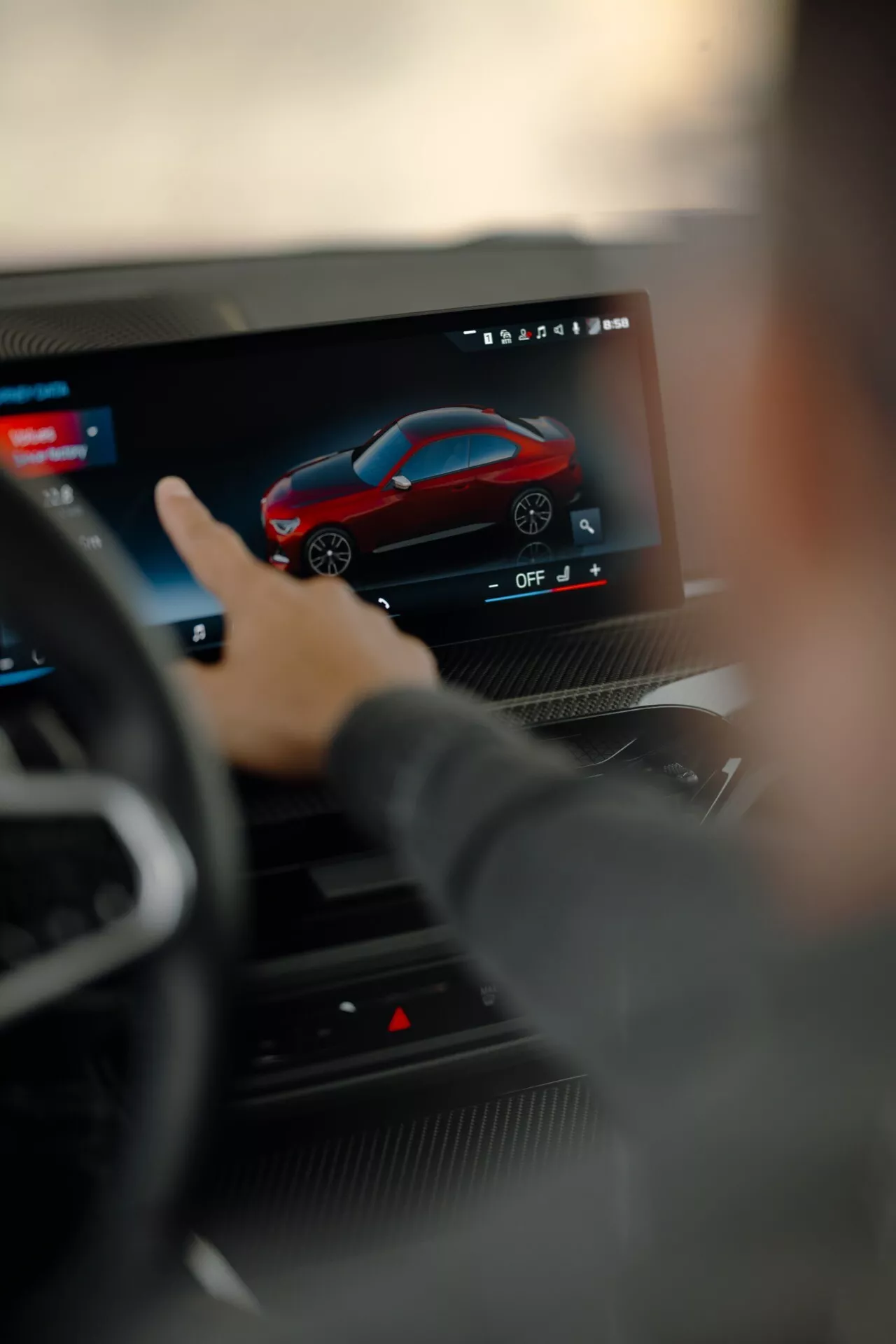 Detailný pohľad na moderný dotykový infotainmentový systém v aute, ako ho vidí vodič, zameriavajúci sa na rozhranie prispôsobenia zobrazujúce červené športové auto. Volant je trochu rozmazaný, zdôrazňujúc vyspelý technologický systém faceliftu BMW 2er radu 2024 G42-G87 s pokročilými funkciami pre navigáciu, nastavenie, konektivitu a multimediálnu zábavu.
