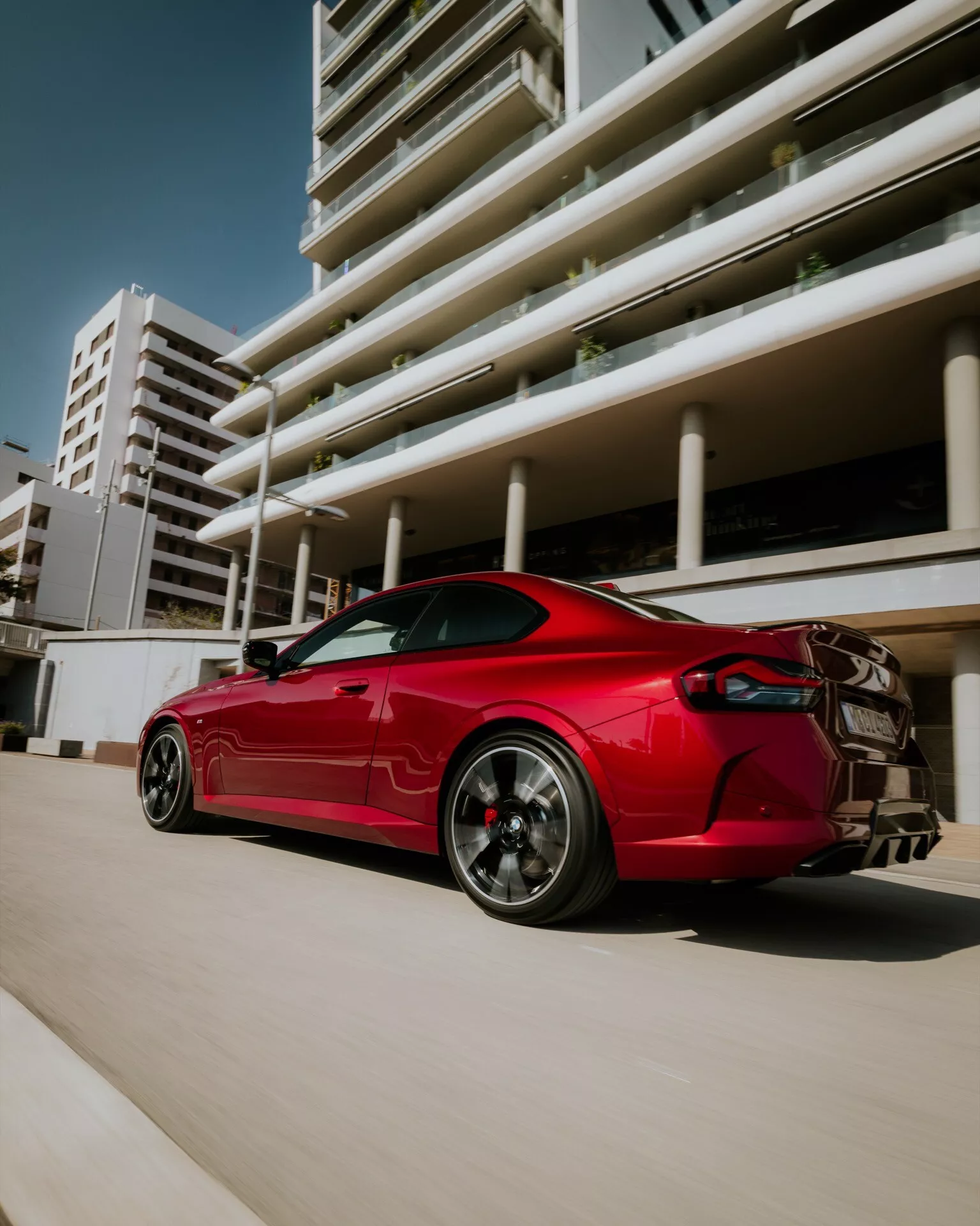 Fotografia modelu BMW radu 2 2024 (facelift G42-G87) v žiarivej červenej farbe, ukazujúca elegantné športové kupé z nízko položeného zadného trojštvrťového pohľadu. Obsahuje aerodynamické línie, grafitové viaclúčové zliatinové kolesá s viditeľnými výkonnostnými brzdovými strmeňmi, výrazný zadný spojler, dvojitý výfukový systém a ostré LED zadné svetlá. Obrázok zachytáva auto v mestskom prostredí s modernými budovami v pozadí, zdôrazňujúci jeho agresívny postoj a športový dizajn.