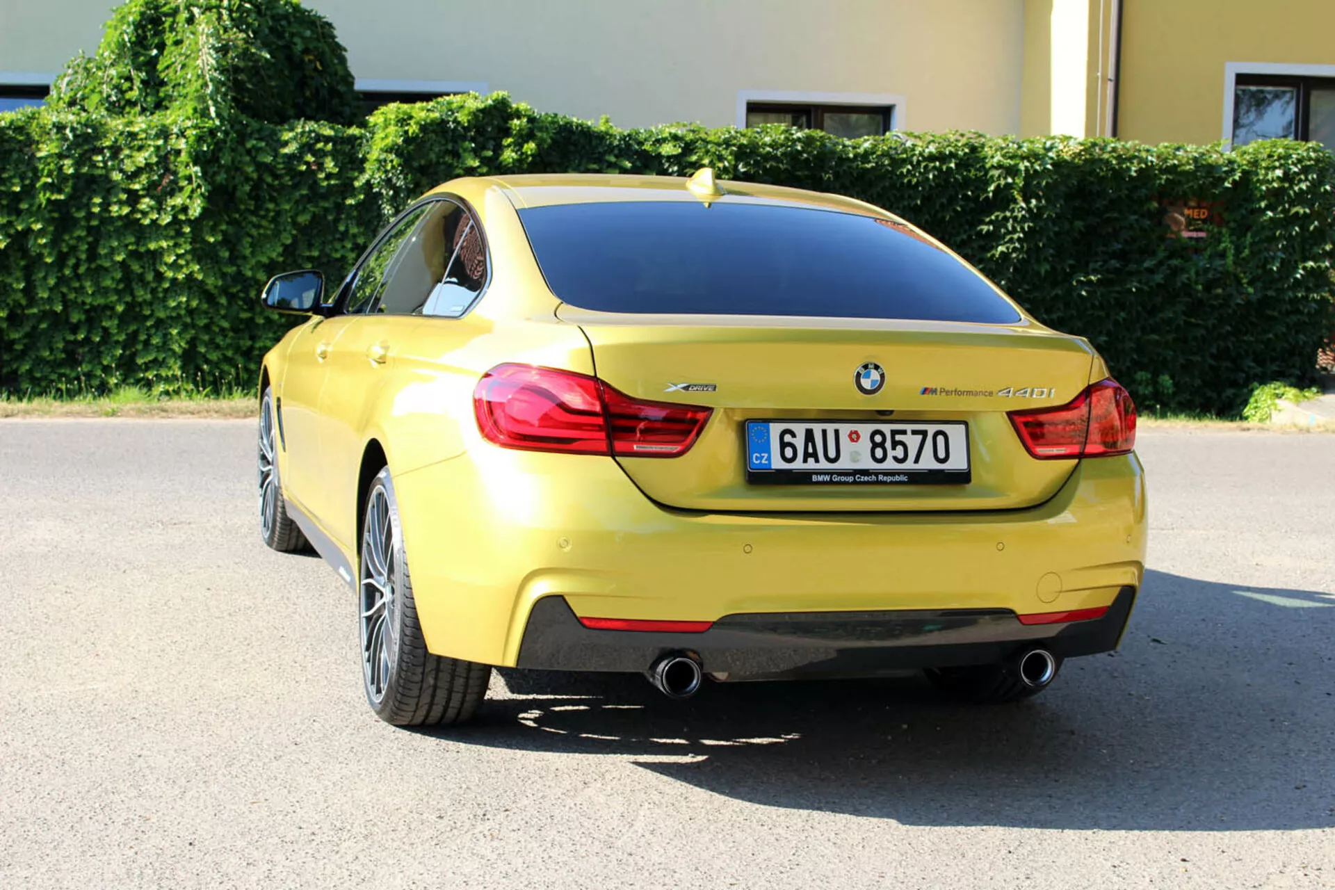 Žlté kupé BMW radu 4 z roku 2013 z pohľadu zozadu pod trojštvrtečným uhlom zobrazuje dizajn batožinového priestoru, zadný nárazník, časť zadného kolesového oblúka, charakteristické zadné svetlá, dvojité výfukové potrubie, evidenčnú značku, logo BMW, označenie modelu, parkovacie senzory, anténu vo forme žraločej plutvy a diskové kolesá z ľahkých zliatin s viacerými paprskami, zaparkované v mestskom prostredí na slnečný deň.