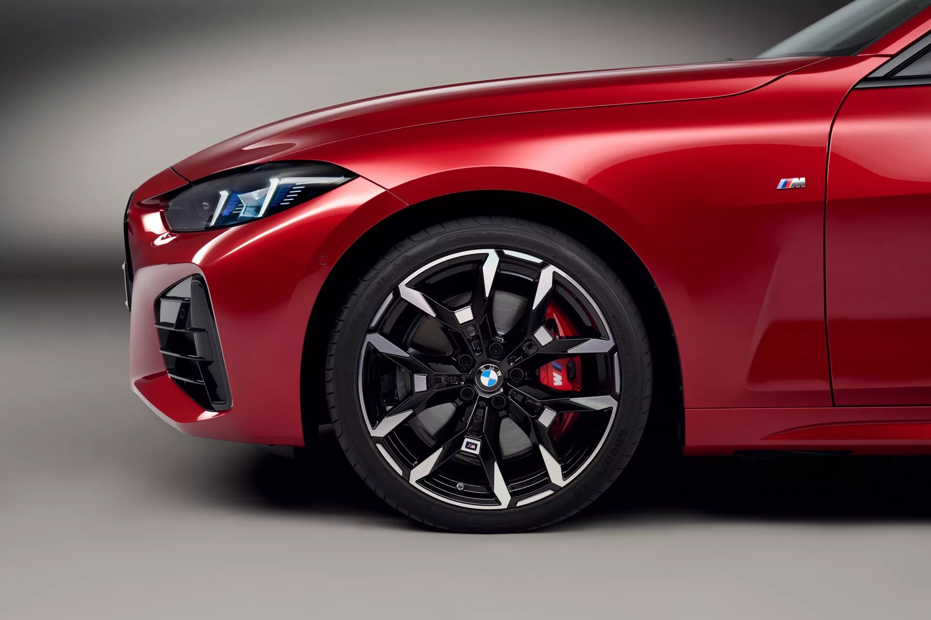 Detailný záber červeného predného štvrťroku BMW radu 4 (G22-G23-G26-G82-G83 facelift) na rok 2024, ukazujúci koleso s viacerými paprskami, výkonnú brzdovú strmene, štíhlé LED svetlomet a odznak 'M', ktorý naznačuje vlastnosti radu BMW M.