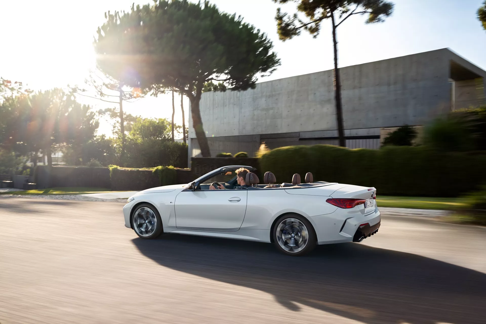 BMW radu 4 Cabrio (G22-G23-G26-G82-G83 Facelift) 2024 v pohybe, predvádzajúce svoj biely elegantný dizajn s výraznými oblúkmi kolies, štýlovými zliatinovými kolesami a čistým výhľadom do jeho luxusného interiéru prostredníctvom mechanizmu sklopné strechy, počas zlatej hodiny s dynamickým, športovým postojem zdôrazneným rozostrením rýchlosti a slnečným žiarom.