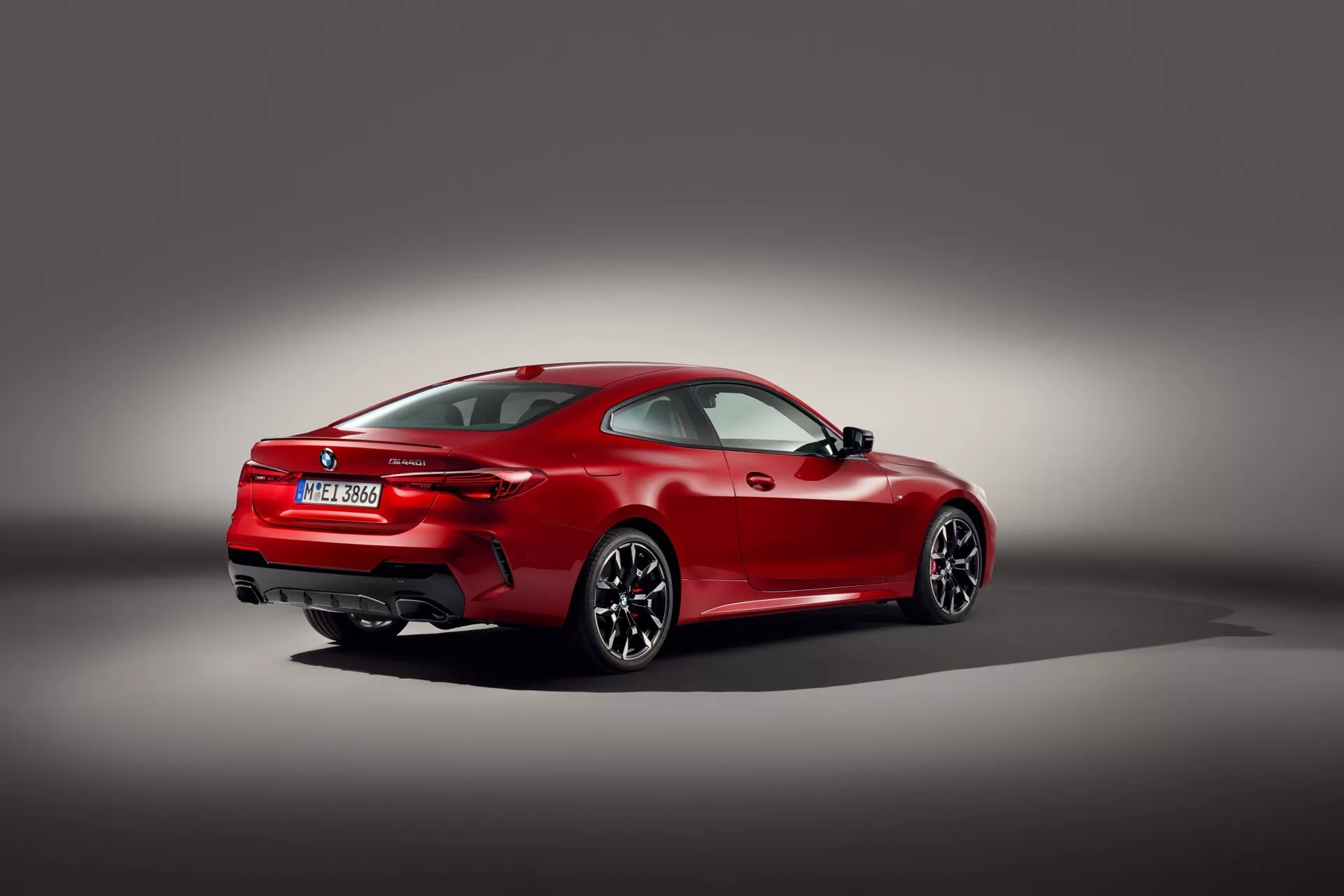 Červené športové kupé BMW radu 4 (G22-G23-G26-G82-G83 Facelift) z roku 2024 v trojštvrťovom zadnom pohľade proti pozadiu so šedým gradientom, predvádzajúce jeho aerodynamický dizajn, svalnatý zadný oblúk kolesa, viaceré lúčové zliatinové kolesá, tenké horizontálne zadné svetlá, dvojité výfukové špičky, integrovaný zadný spojler a LED technológiu na pneumatikách s nízkym profilom pre vysoký výkon, odzrkadľujúce zameranie na luxus a vysoký výkon.