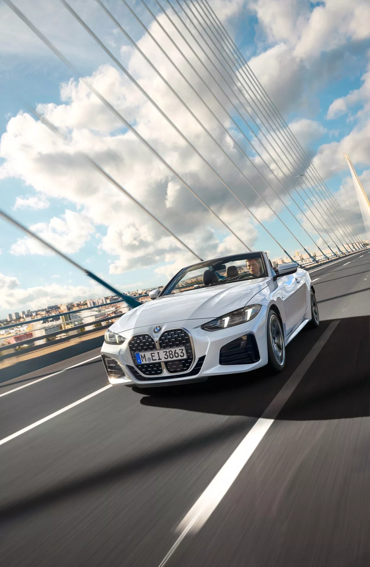 Biely kabriolet BMW radu 4 ročník 2024 (facelift G22-G23-G26-G82-G83) dynamicky zachytený z nízkeho uhla spredu pri jazde po závesnom moste pod čiastočne zamračenou oblohou, predvádzajúci svoj elegantný dizajn, LED osvetlenie a aerodynamické vlastnosti