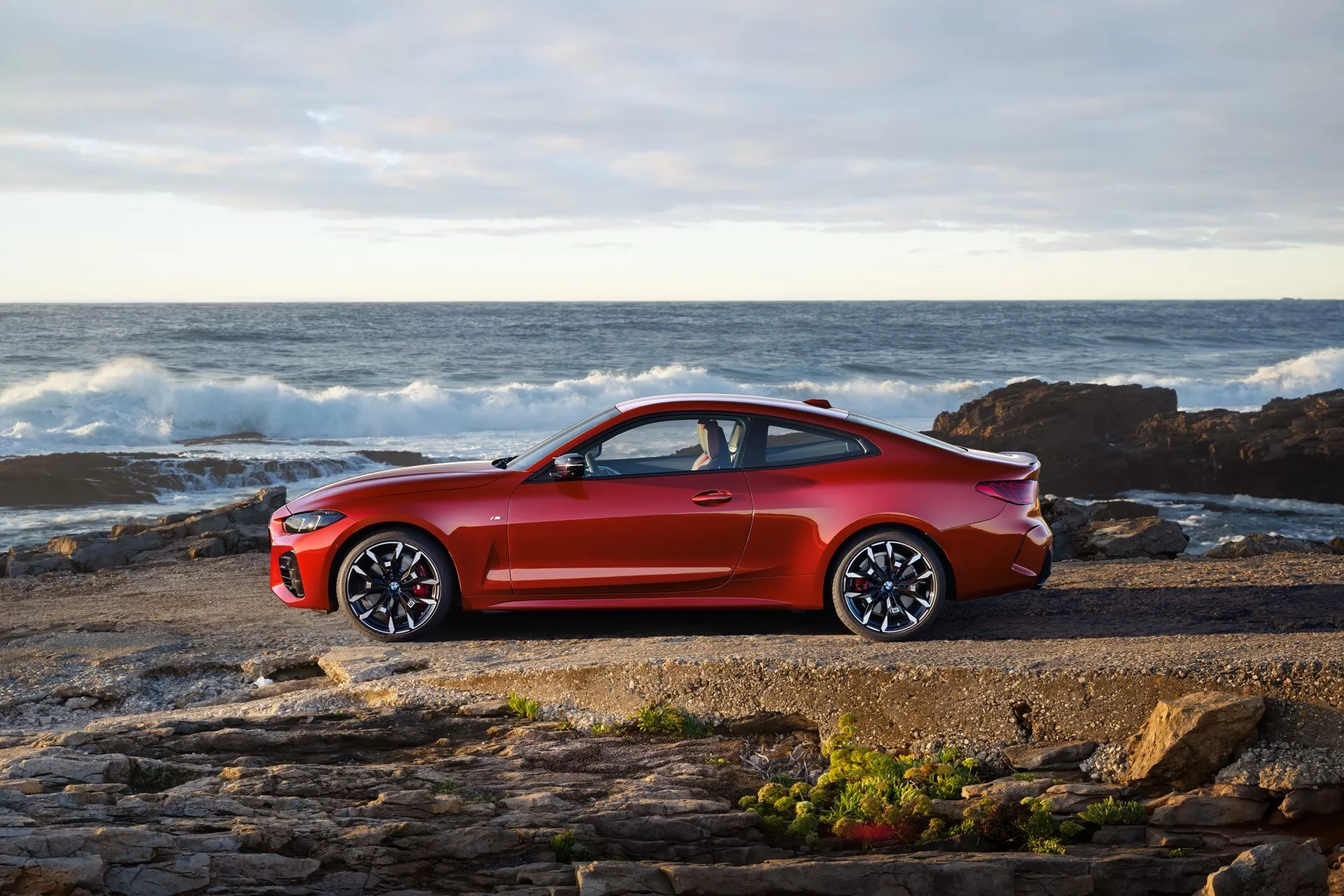 Coupe BMW radu 4 (G22-G23-G26-G82-G83 Facelift) z roku 2024 v jasne metalickej červenej farbe zaparkované pri skalnatom brehu s vlnami oceánu na pozadí pod čiastočne zamračenou oblohou, ktoré ukazuje svoj športový bočný profil s veľkými viaceramennými zliatinovými kolesami a elegantným aerodynamickým dizajnom