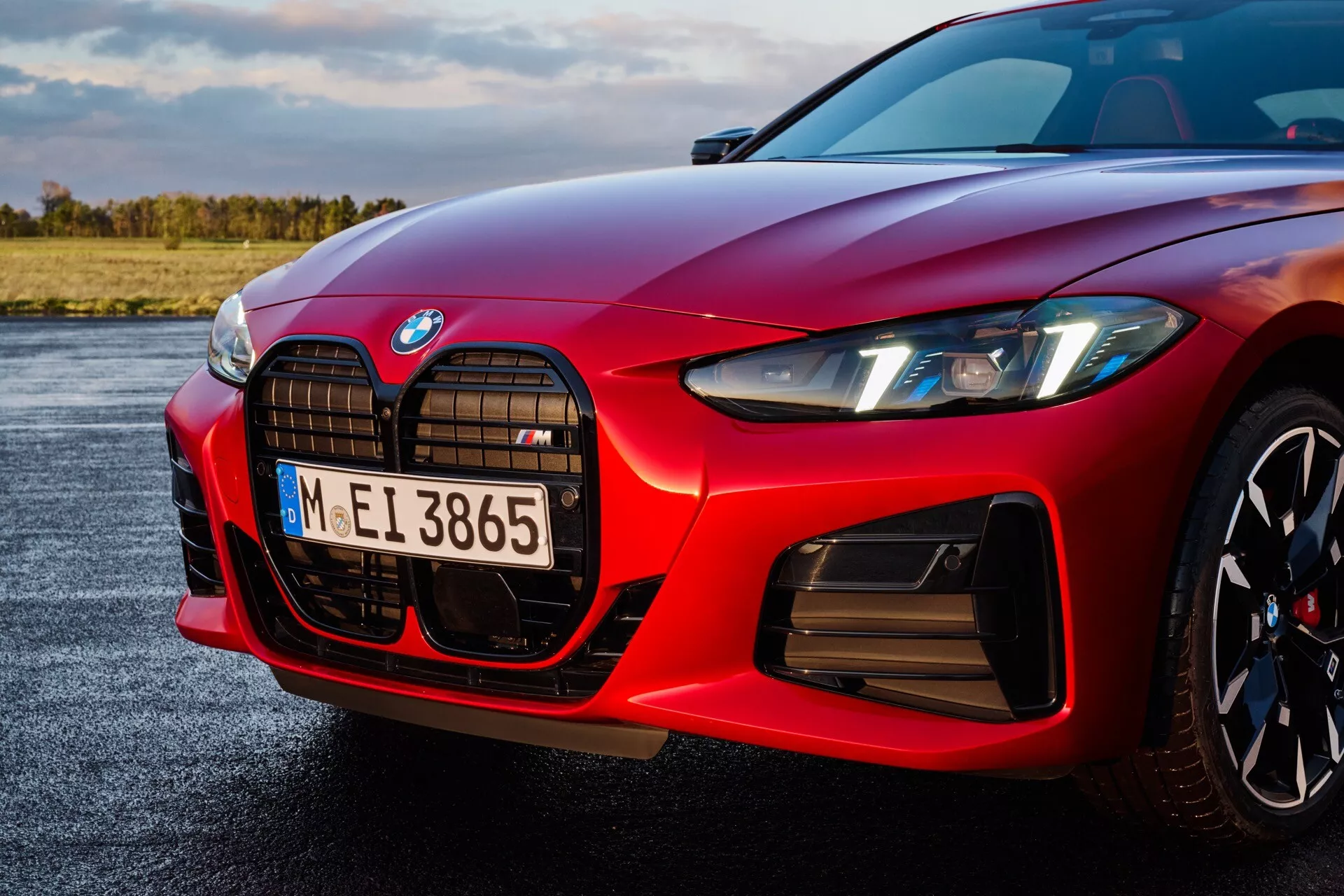 Detailný pohľad na prednú ľavú stranu červeného BMW radu 4 2024 model (G22-G23-G26-G82-G83 facelift), zvýrazňujúci charakteristickú vertikálne orientovanú obličkovitú mriežku s centrálnym emblémom, ostré LED predné svetlo s dennými svetlami, konturovaný dizajn kapoty smerujúci k mriežke pre lepšiu aerodynamiku, predný nárazník s veľkými vzduchovými vstupmi naznačujúcimi pokročilé chladenie pre výkon a odznak 'M', ktorý označuje vysokovýkonnú variantu, proti lesklému červenému povrchu.
