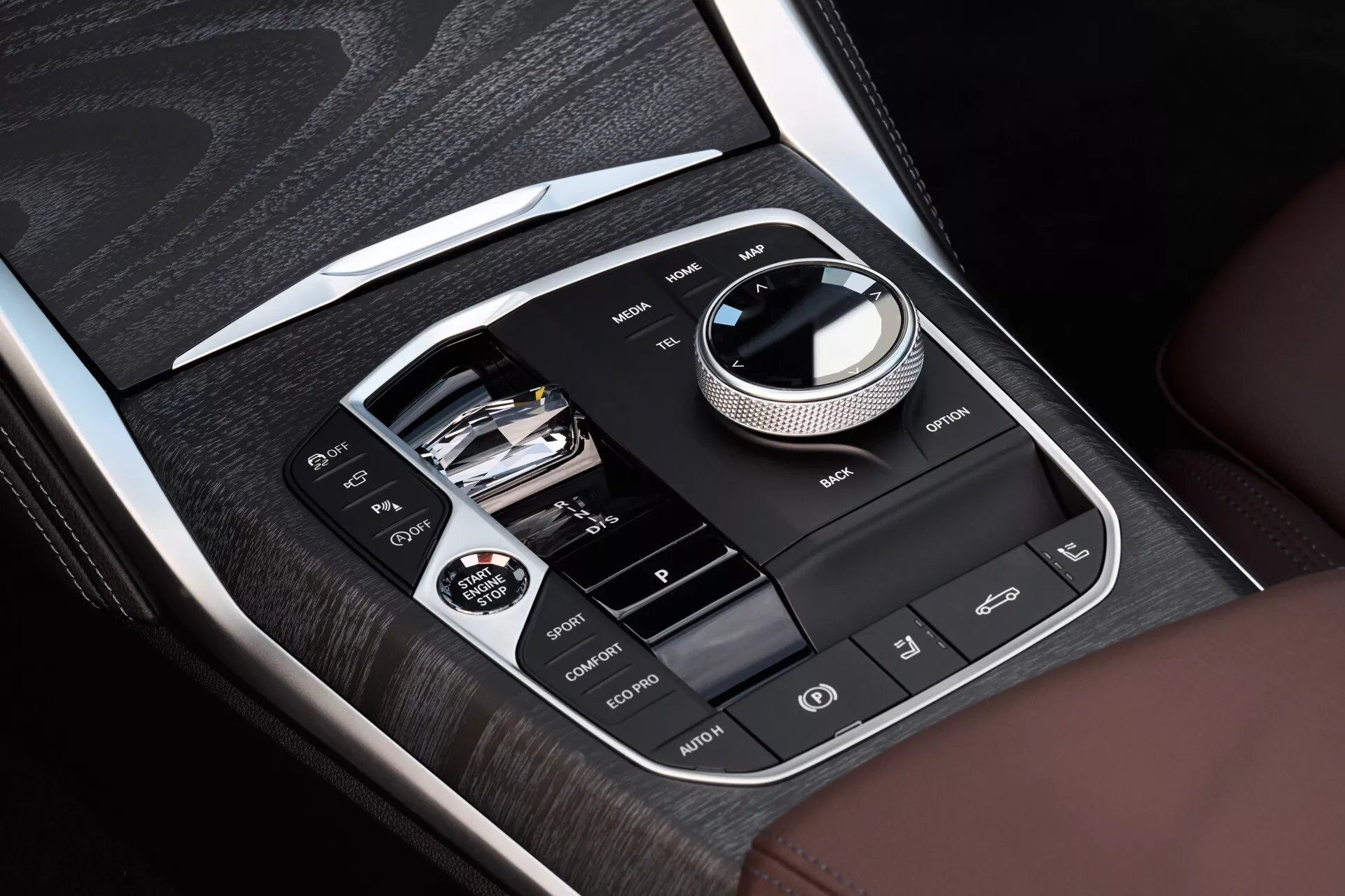 Detailný pohľad na centrálnu konzolu v BMW radu 4 z roku 2024 (facelift G22-G23-G26-G82-G83) zobrazujúci informačné a ovládacie systémy s drevodekorom a chrómovými akcentmi, s hlavným kruhovým rozhraním, konektivitou USB a slotom pre SD kartu, a tlačidlá pre asistenciu vodiča vrátane vyhrievania sedadiel a vypnutia systému ESC