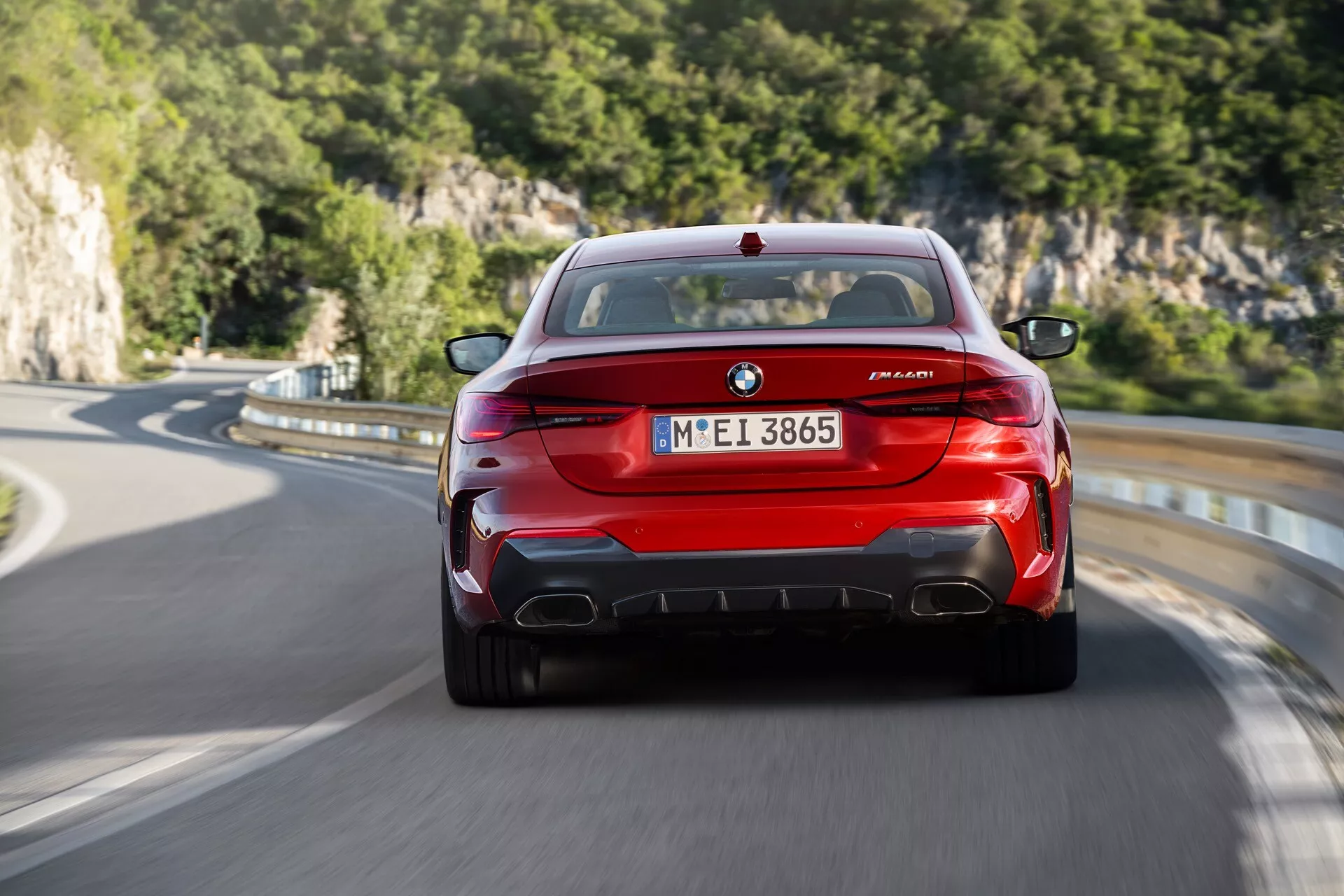 Zadný pohľad na červené BMW M8 Competition v pohybe, predvádzajúce emblém BMW, označenie M8 a odznak série M, unikátne zadné svetlá, štvornásobné výfukové rúry a aerodynamický zadný difúzor na vidieckej ceste s prírodným pozadím.