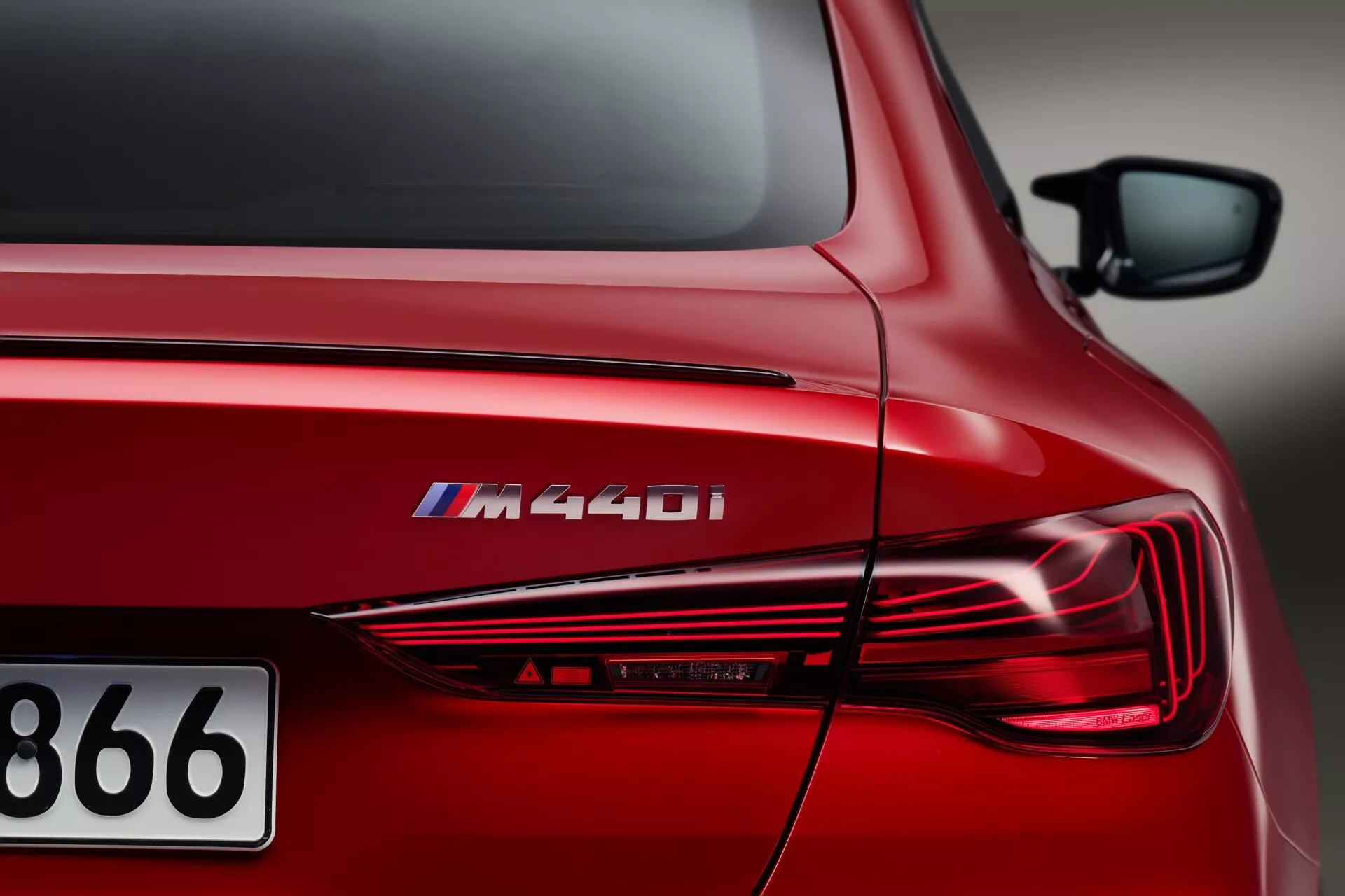 Detailný šikmý pohľad na zadnú štvrtinu červeného BMW radu 4 (G22-G23-G26-G82-G83-Facelift) M440i, zobrazujúci označenie modelu, štýlové LED zadné svetlá, sochársky spracovaný vonkajší dizajn a lesklý metalický červený povrch pri umelom osvetlení.