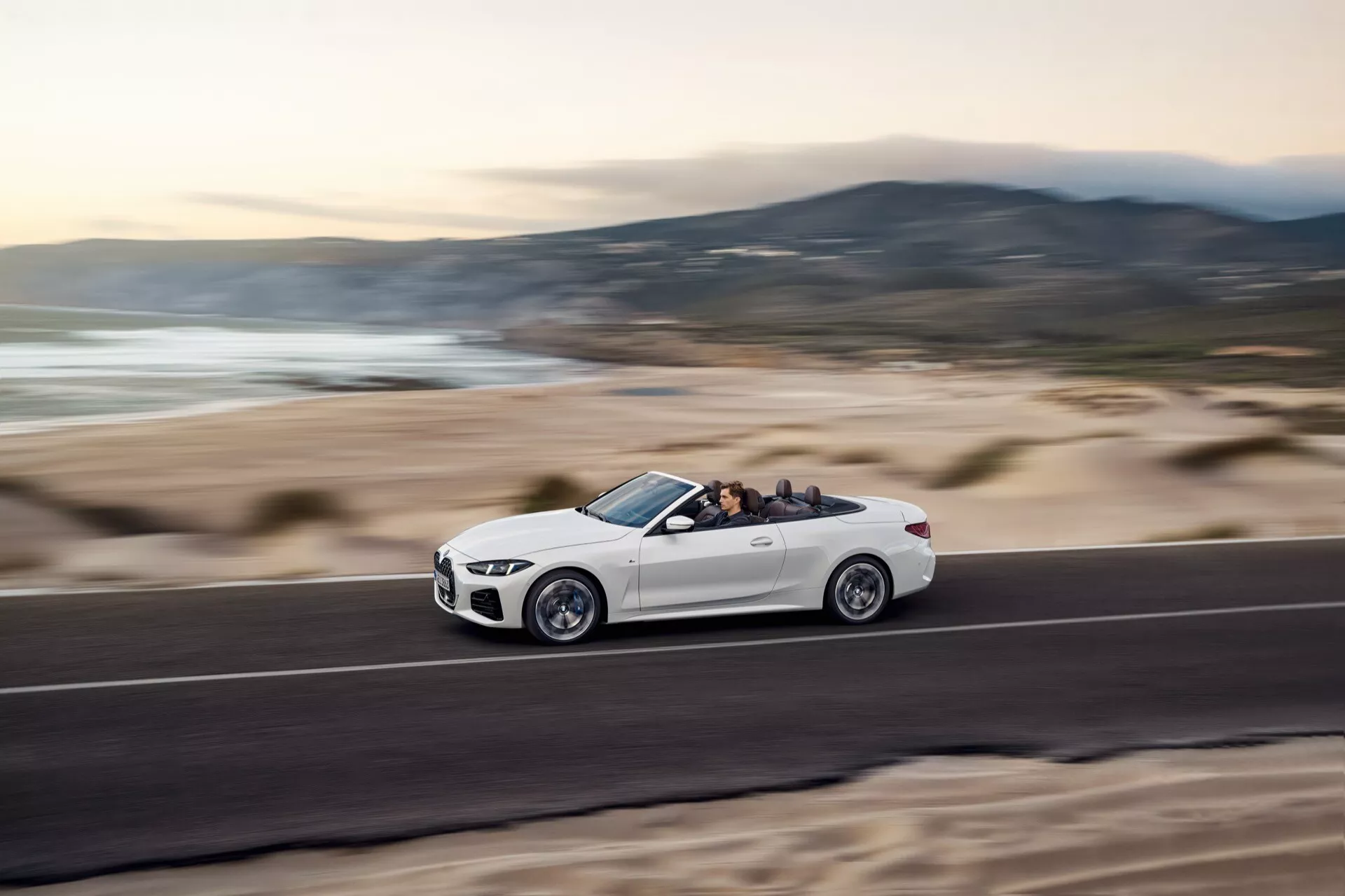 BMW radu 4 kabriolet 2024 (G22-G23-G26-G82-G83 Facelift) v pohybe na pobrežnej ceste, zobrazuje bočný profil s dynamickým postojom vďaka snímke z nízkeho uhla, viditeľné detaily kolies a výbavy, zložená strecha odhaľuje interiér a obsadenie, proti rozostrenému pozadiu plážovej krajiny