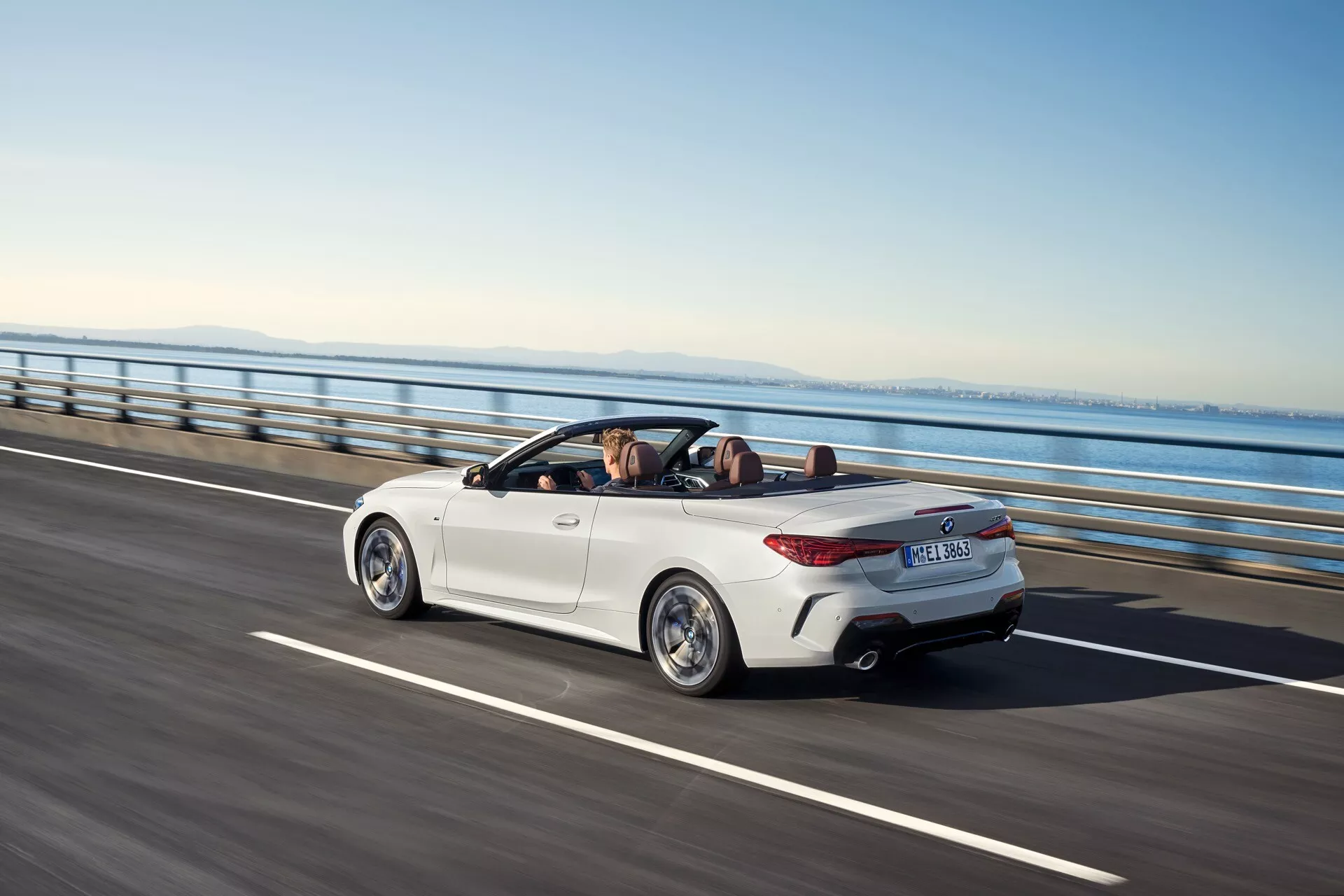 Kabriolet BMW radu 4 (G22-G23-G26-G82-G83 Facelift) rok 2024 v jasne bielej farbe, prezentujúci svoju sklápateľnú strechu, LED zadné svetlá a zliatinové kolesá z trojštvrťového zadného pohľadu na ceste pod jasným nebom