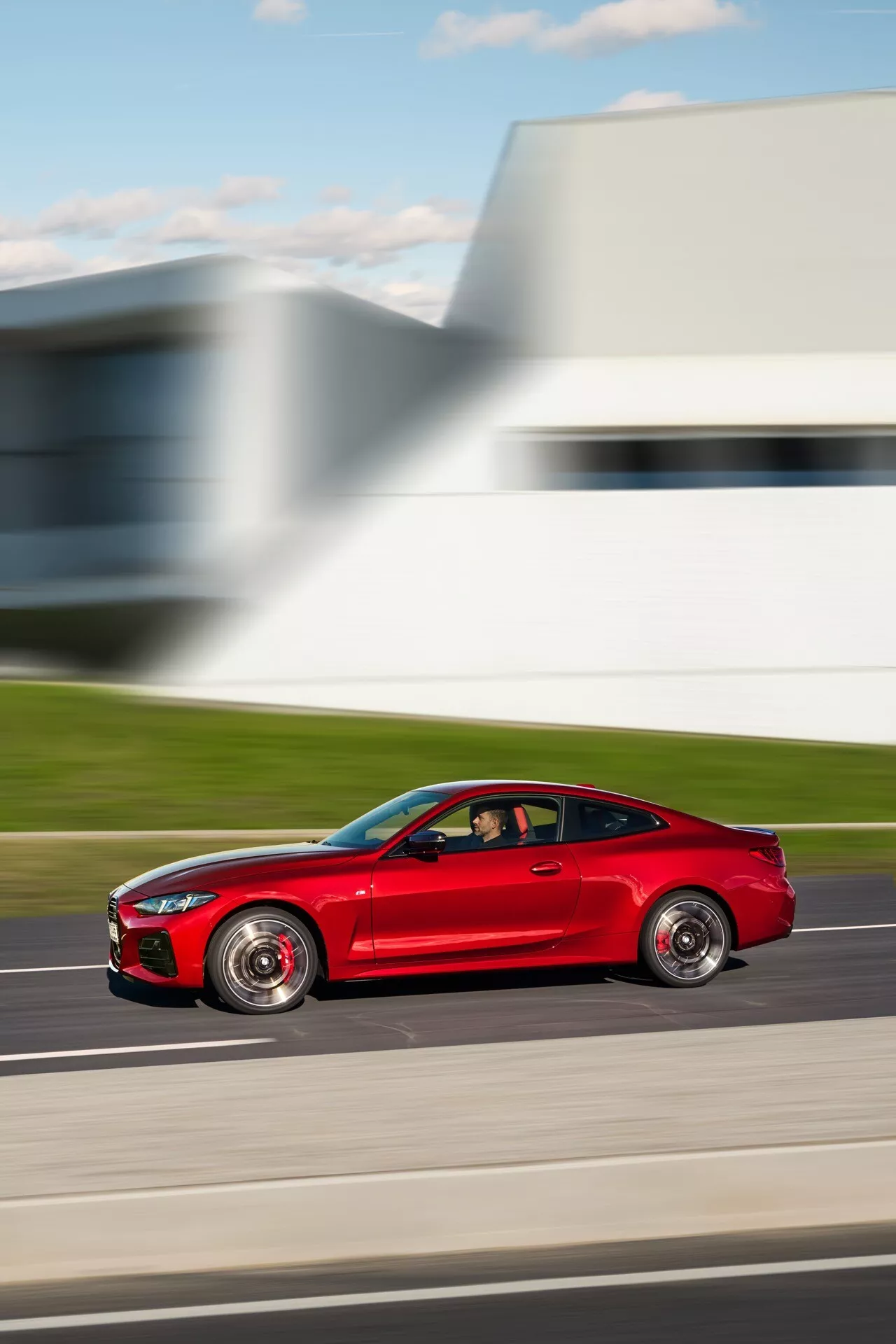 Športové kupé BMW radu 4 pre rok 2024 (facelift G22-G23-G26-G82-G83) v pohybe, červené športové kupé s aerodynamickými bočnými zrkadlami, výraznými diskami a nízkoprofilovými pneumatikami, zachytené z bočného pohľadu, ktorý zdôrazňuje dynamický dizajn a rýchlosť auta proti rozostrenému pozadiu moderného architektúry.