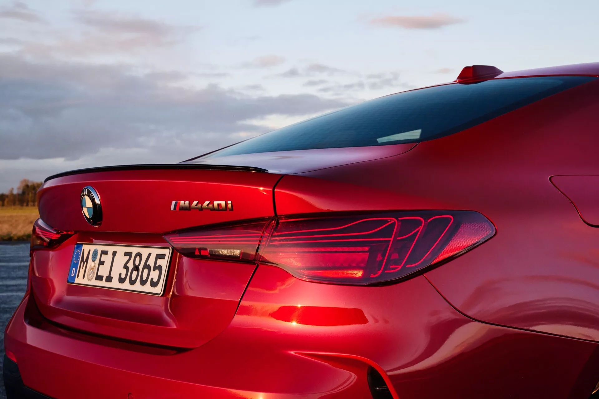 Zadná časť BMW série 4 2024 (G22-G23-G26-G82-G83 Facelift) zobrazená z vysokého trojštvrťového uhla, predstavujúca pravý zadný kvartál, dizajn LED zadných svetiel, integrovaný zadný difúzor, časť zadného okna a stĺpika C, so zameraním na prvky dizajnu luxusných športových sedanov, označenie výkonu a metalický červený povrch v exteriéri pri východe alebo západe slnka.