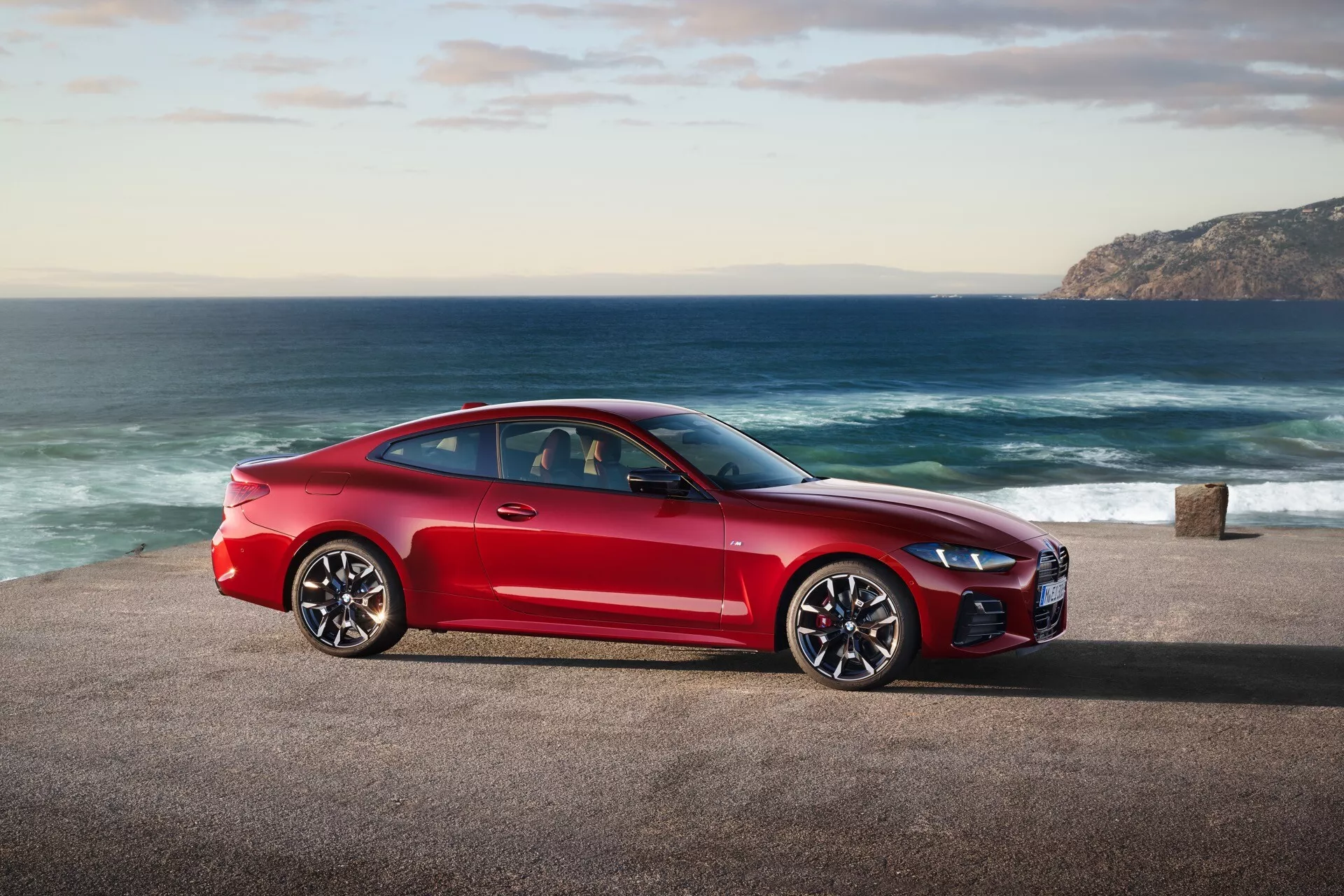 Červené športové kupé BMW série 4 (facelift G22-G23-G26-G82-G83) 2024 zaparkované na pobrežnej ceste s oceánom a kopcovitým pobrežím na pozadí, zobrazujúce profil vozidla zo strany, elegantný dizajn a výkonnostné prvky v prírodnom dennom svetle