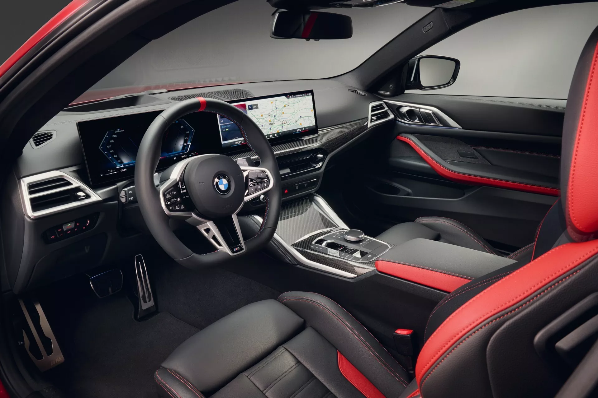 Interiérový pohľad na BMW radu 4 z roku 2024 (G22 G23 G26 G82 G83 Facelift) zobrazujúci stranu vodiča, vrátane palubnej dosky a oblasti volantu s multimediálnymi ovládacími prvkami, digitálnym prístrojovým panelom, širokouhlým displejom infotainment, ergonomickou radiacou pákou, kvalitnými materiálmi s červenými akcentmi a pokročilými technologickými vlastnosťami.