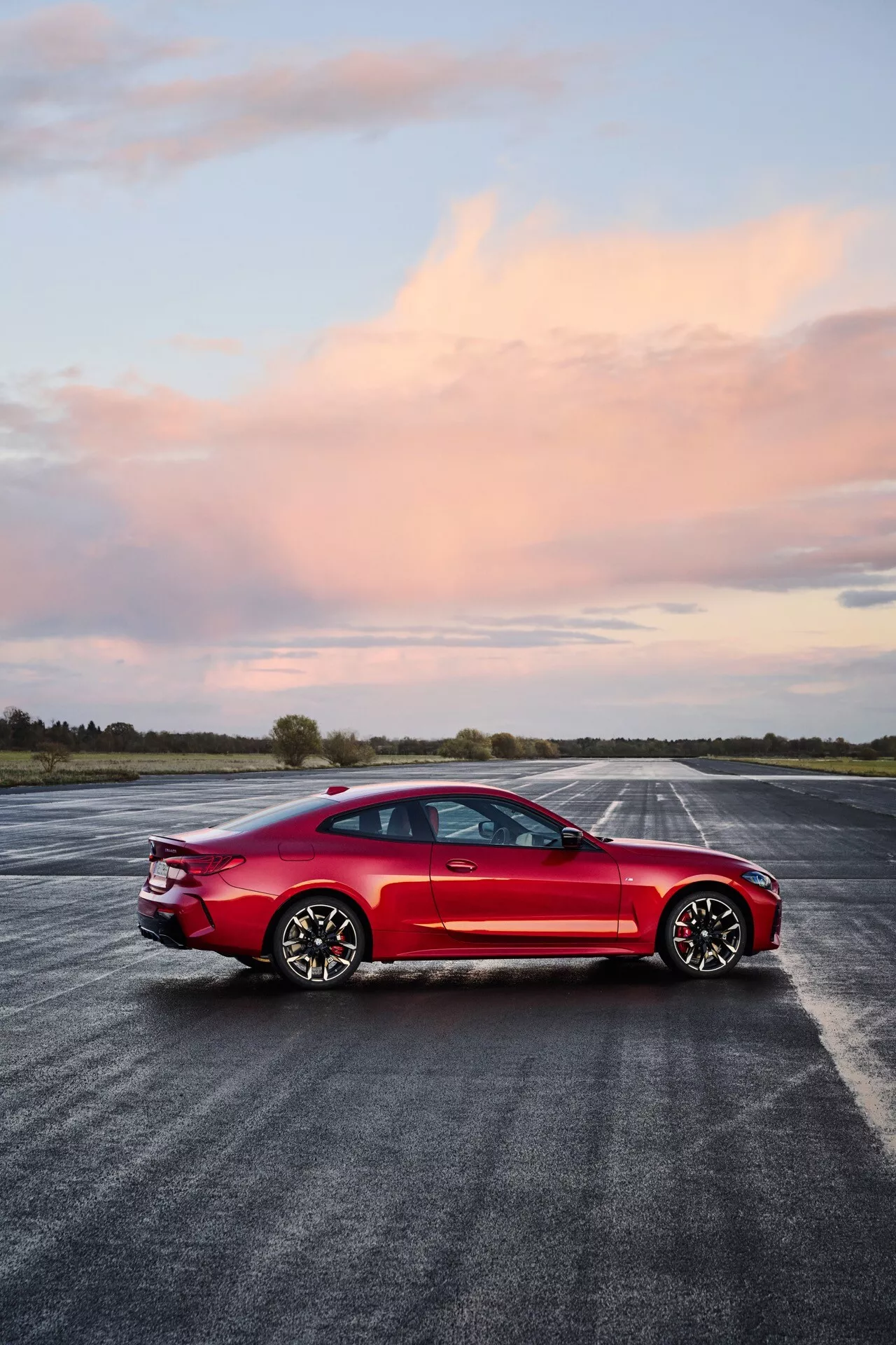 Červený štvordverový športový sedan BMW rady 4 (G22-G23-G26-G82-G83 Facelift) na mokrom asfalte, zobrazujúci bočný profil, aerodynamický dizajn, LED svetlomety, panoramatickú strechu, viaclopatkové zliatinové kolesá a vysokovýkonné brzdy pod večernou oblohou