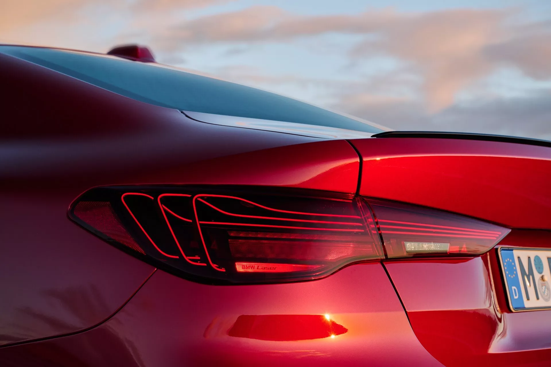Detailný pohľad na ľavé zadné svetlo BMW radu 4 2024 (G22-G23-G26-G82-G83 facelift) s horizontálnymi LED pruhmi a aerodynamickým dizajnom plutiev na pozadí západu slnka