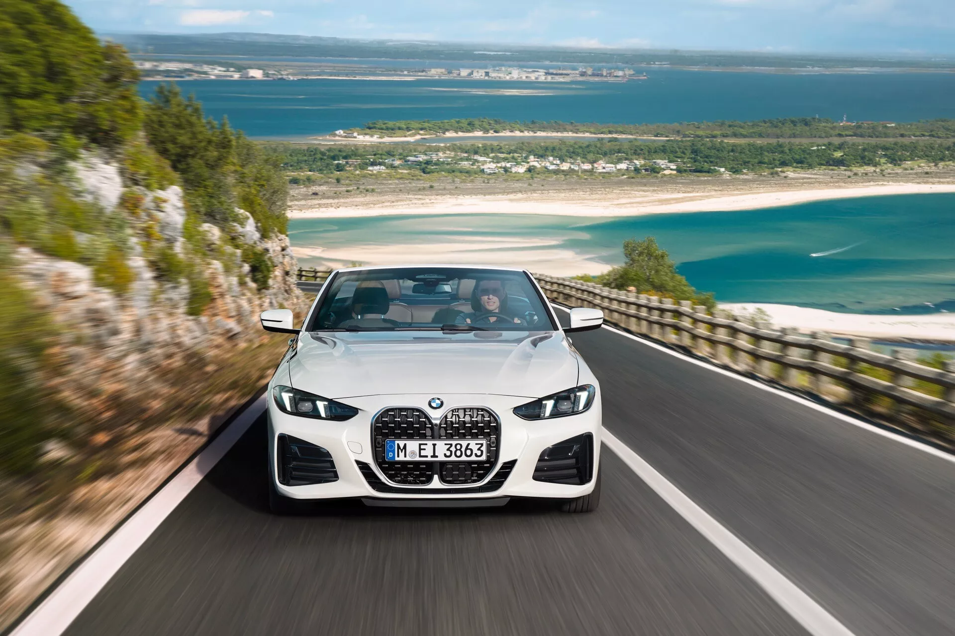 Luxusný sedan BMW radu 4 (G22-G23-G26-G82-G83 facelift) z roku 2024 idúci pozdĺž malebnej pobrežnej cesty, ktorý ukazuje svoju veľkú obličkovitú mriežku, uhlové LED svetlomety a dynamický dizajn predného nárazníka