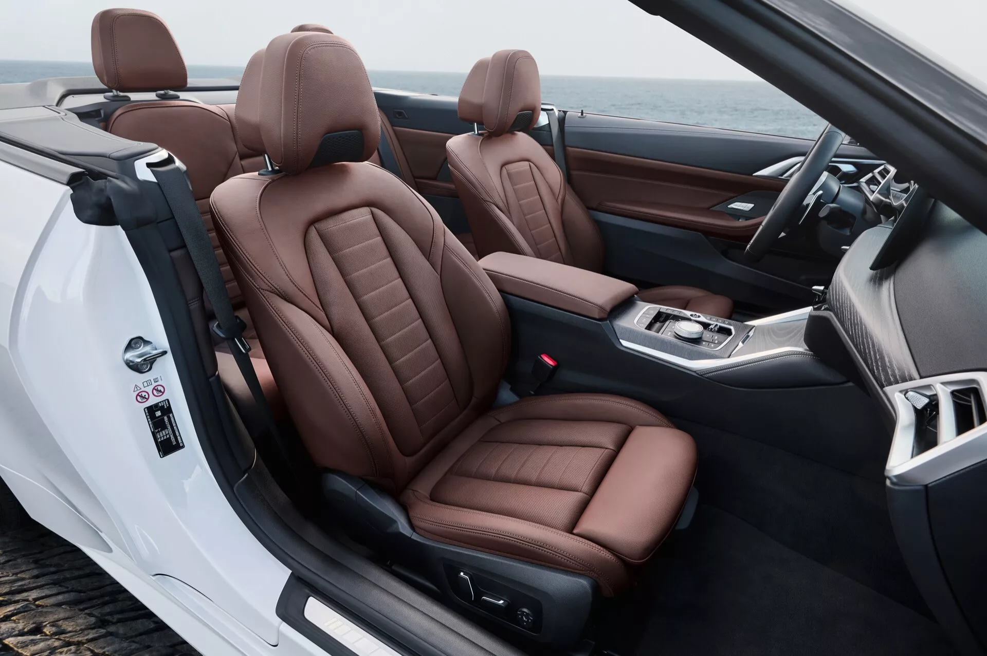 Interiér BMW radu 4 2024 (G22-G23-G26-G82-G83 facelift) so zameraním na prednú kabinu z pohľadu zo zadnej strany, zdôrazňujúci luxusné hnedé kožené sedadlá, kabrioletovú povahu a pokročilé ovládacie prvky na dverách a na stredovej konzole.