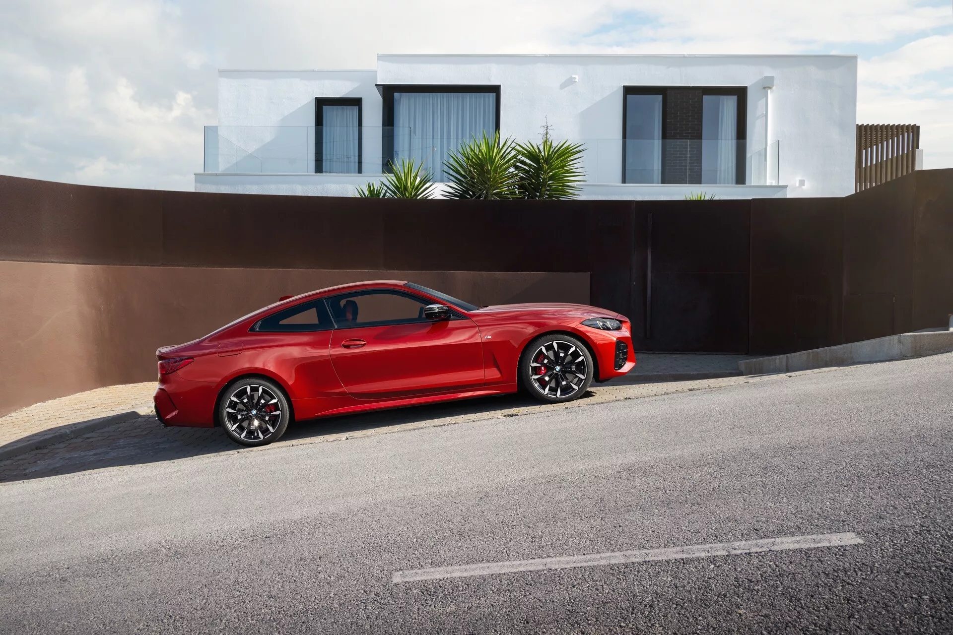 Svetlo červené dvojdverové kupé BMW radu 4 (G22-G23-G26-G82-G83 Facelift) 2024 zaparkované na strane ulice, predvádzajúce svoj štýlový, športový dizajn s veľkou výraznou mriežkou, LED svetlometmi, viacerými hovorovými zliatinovými kolesami, pneumatikami s nízkym profilom, vysoce kvalitnou lesklou povrchovou úpravou, integrovanými smerovkami na bočných zrkadlách a vysoko výkonným brzdovým systémom v obytnej oblasti.