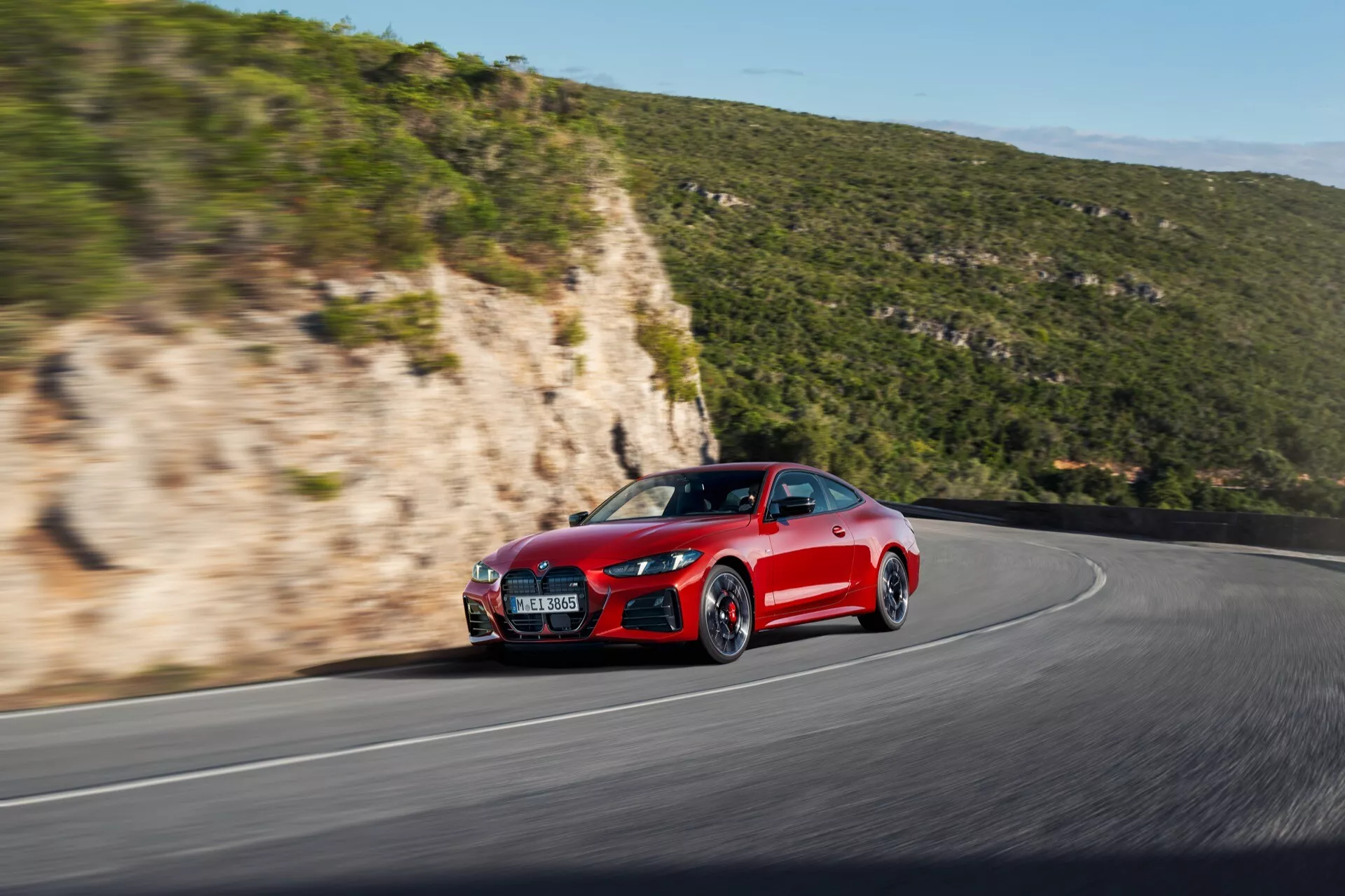 Dynamický bočný predný pohľad na červené športové auto BMW radu 4 2024 (G22-G23-G26-G82-G83 facelift) ukazujúce jeho agresívny štýl, aerodynamické linky, športový predný nárazník, kapotu, bočný panel, predné koleso a pneumatiku na kľukatej ceste