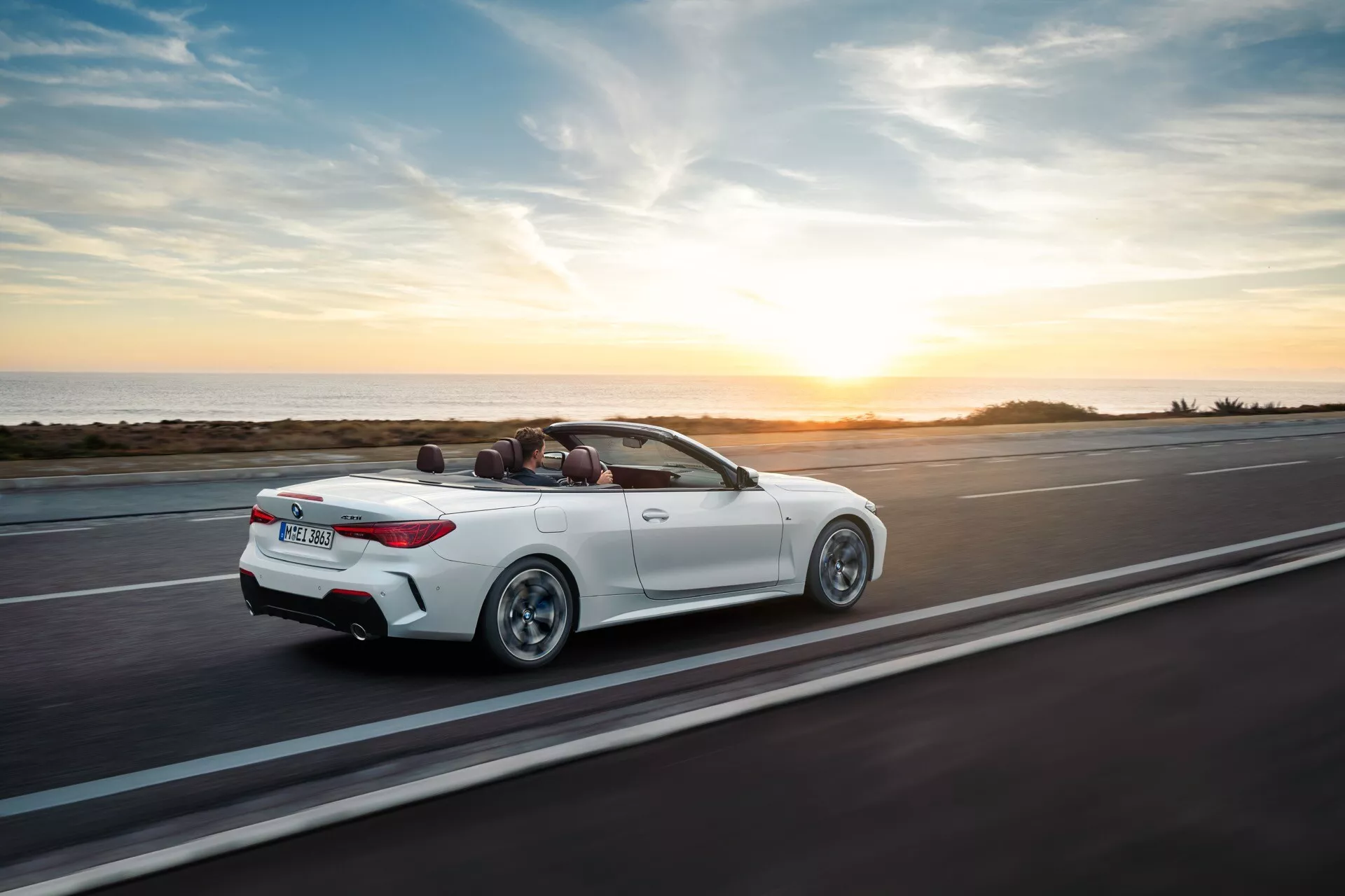 BMW radu 4 kabriolet (facelift G22-G23-G26-G82-G83) roku 2024 v pohybe na pobrežnej ceste pri západe slnka, pohľad zozadu a zhora ukazujúci jeho bielu vonkajšiu stranu, sklopenú strechu kabrioletu a športový dizajn