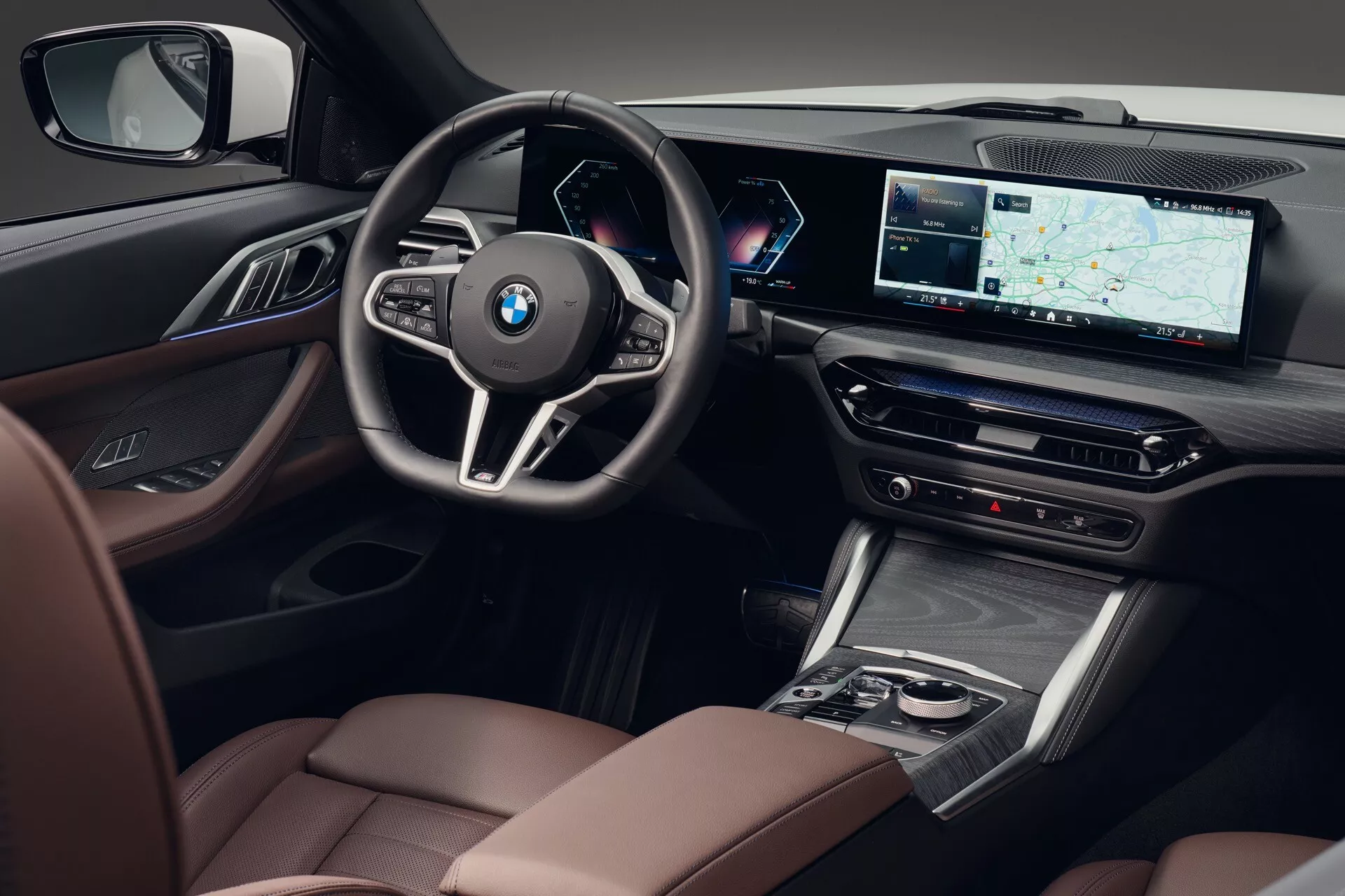 Interiérový záber z mierne zvýšeného uhla zameraný na stranu vodiča BMW série 4 2024 (G22-G23-G26-G82-G83 facelift), predvádzajúci sedadlá s kvalitnou koženou čalúnením s kontrastným prešívaním, drevené alebo drevené efekty, ovládacie prvky prémiového audio systému na dverovom paneli, športový trojramenný multifunkčný volant s emblémom BMW, úplne digitálny a prispôsobiteľný prístrojový panel za volantom, veľký širokouhlý displej infotainmentu s GPS navigáciou, ovládanie klímy, elegantnú radiacu páku, stredovú konzolu s dotykovým panelom alebo ovládačom obklopeným chrómovými detailmi a čiastočne viditeľné luxusné sedadlo spolujazdca, zdôrazňujúce technologické a luxusné funkcie.