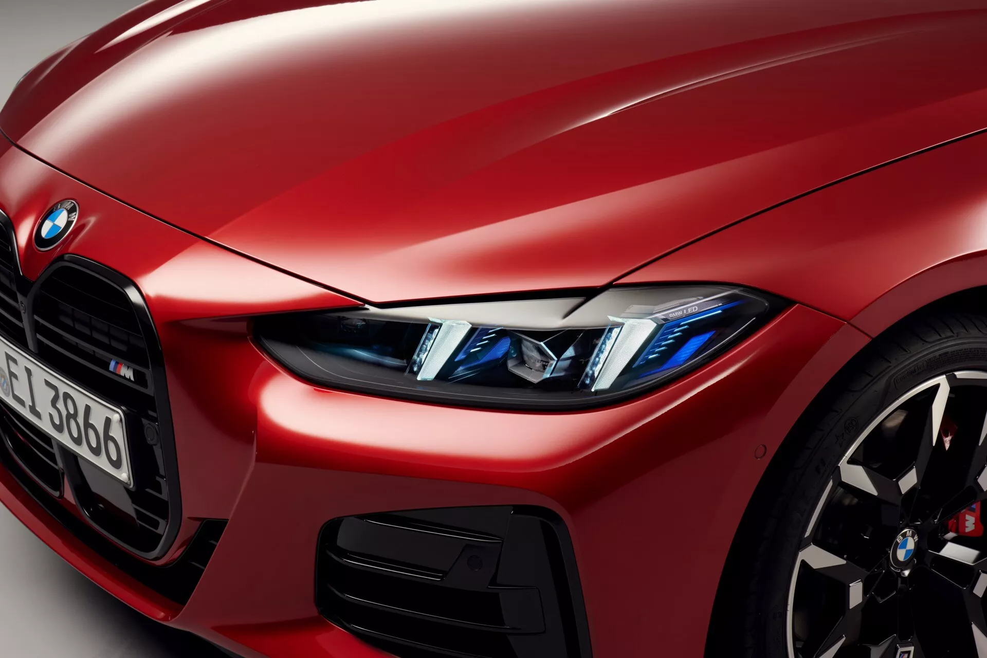 Detailný pohľad na červené BMW radu 4 z roku 2024 (facelift G22-G23-G26-G82-G83), zobrazuje prednú ľavú štvrtinu s dôrazom na LED svetlomety s anjelskými očami, pohľad na ikonickú obličkovú mriežku, športové zliatinové koleso s vysokovýkonnými brzdami, metalický lak a karosériu s detailom prívodu vzduchu a čiastočne viditeľným emblémom BMW a identifikátorom série.