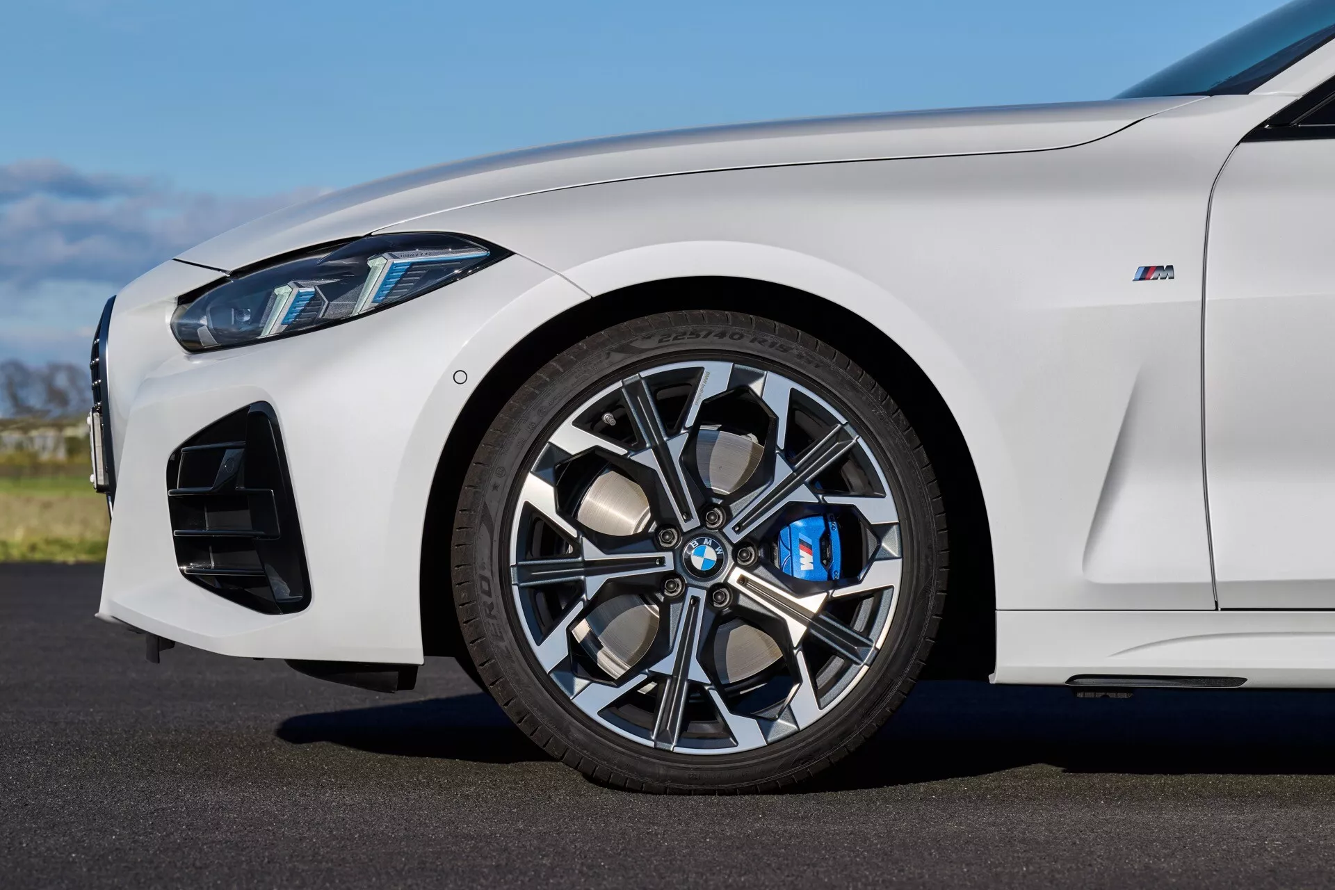 Podrobný bočný pohľad na biely BMW rady 4 (G22-G23-G26-G82-G83 Facelift) 2024 so zameraním na predné koleso s viacerými hovormi zliatinového disku, modrým brzdovým strmene a výkonnostne orientovanými vzduchovými vstupmi pri prirodzenom dennom svetle