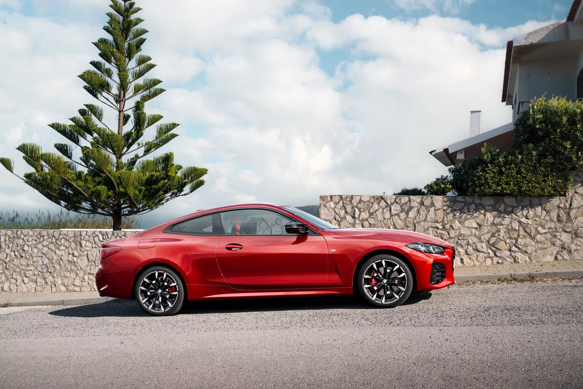 Červené športové kupé BMW radu 4 2024 (G22-G23-G26-G82-G83-Facelift) zaparkované pri obrubníku, ukazujúce celý svoj bočný profil s dynamickou perspektívou z nízkeho uhla zdôrazňujúcou jeho športový dizajn, dlhú kapotu, štíhlu líniu strechy, veľké zliatinové kolesá, aerodynamické bočné zrkadlá, zapustené kľučky dverí, vysoko výkonný brzdový systém s červenými strmeňmi, LED svetlomety a agresívny predný nárazník, pod čiastočne zamračenou oblohou vedľa zelene a rezidenčnej budovy