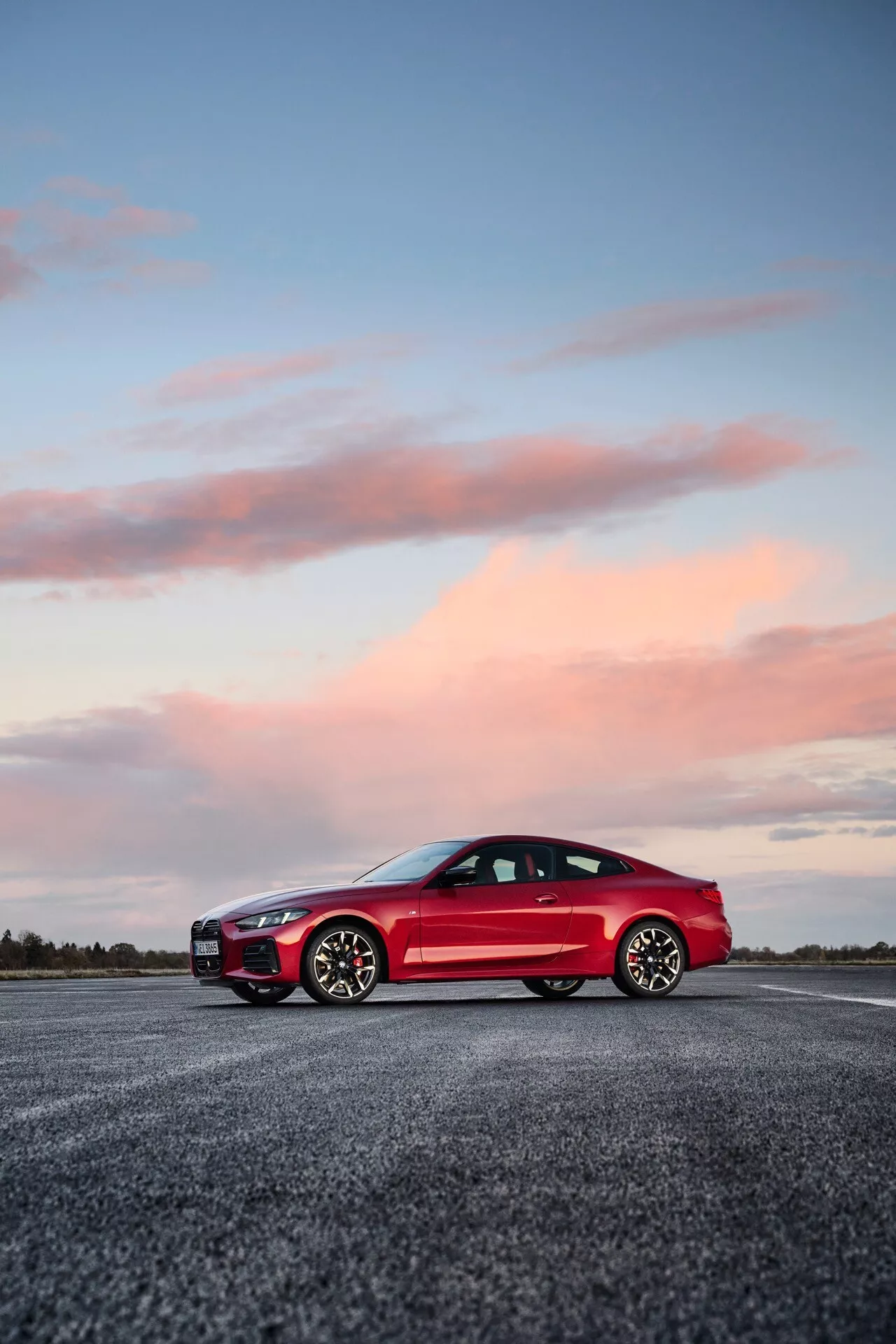 Červené športové auto s dvomi dverami BMW série 4 2024 (G22-G23-G26-G82-G83 facelift) zvýraznené z nízkeho uhla, čím sa zdôrazňuje jeho postoj, dizajnové línie a technologické prvky, ako sú LED svetlomety a aerodynamické zrkadlá, na pozadí súmraku