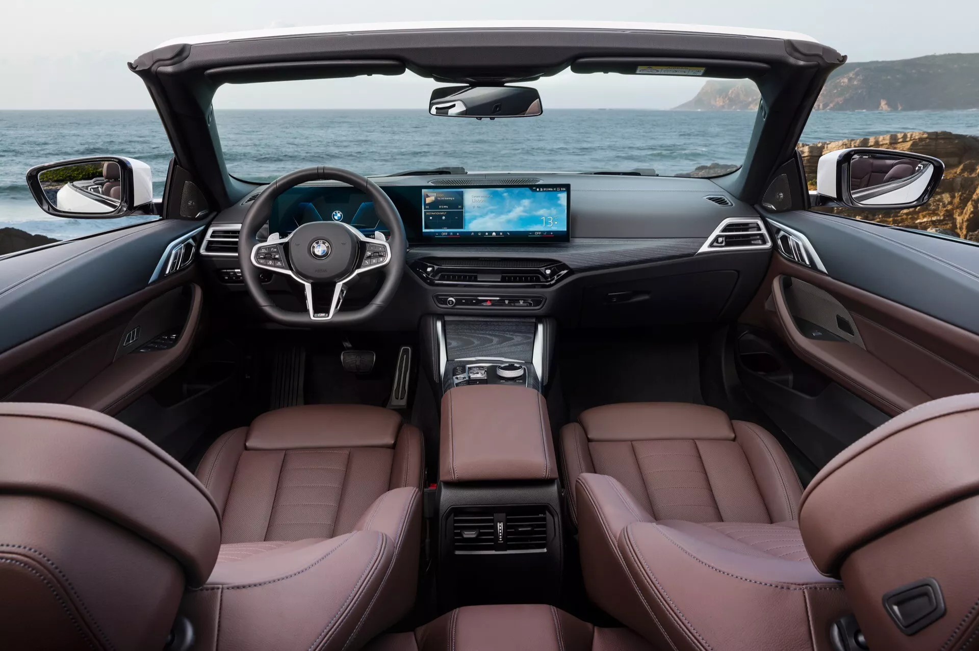 Interiér kabrioletu BMW radu 4 pre rok 2024 s hnedými koženými prednými sedadlami, volantom s tromi ramenami a integrovanými ovládacími prvkami, digitálnym širokouhlým displejom palubnej dosky a elegantnou stredovou konzolou uprostred luxusnej kabíny s odobratou strechou, zobrazujúci scenický pobrežný výhľad