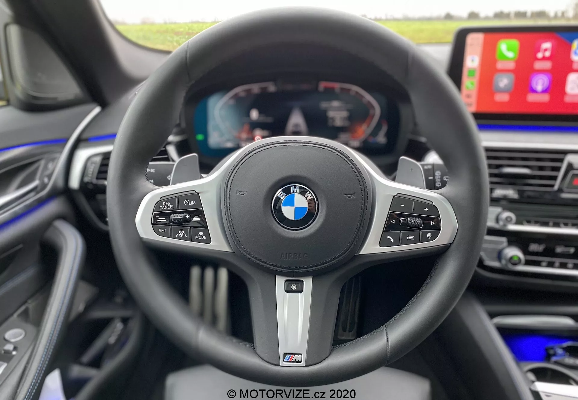Pohľad na interiér z perspektívy vodiča v interiéri BMW 520i Touring Steptronic z roku 2020, zvýrazňujúci športový trojramenný volant s odznakmi BMW a M. Volant obsahuje niekoľko ovládacích tlačidiel pre tempomat, médiá, hlasové povely a telefónne funkcie, a pádla na radenie naznačujúce automatickú prevodovku s manuálnymi možnosťami. Za volantom je plne digitálny displej zobrazujúci tachometer, otáčkomer a informácie o vozidle. Na pravej strane je viditeľná časť stredovej konzoly s veľkým ekranom infotainmentu s funkciou Apple CarPlay, zdôrazňujúc luxus, pokročilé technológie a dizajn zameraný na vodiča.
