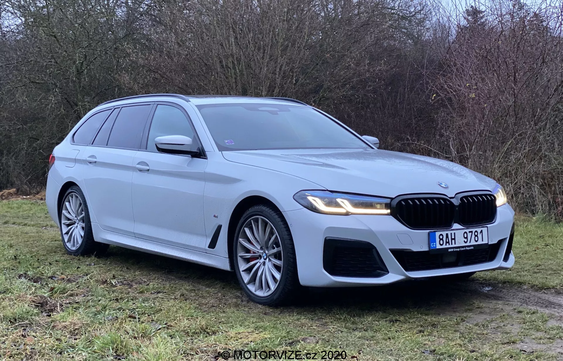 Strieborný BMW 5 Series Touring 2020 zaparkovaný na tráve, viditeľný z trojštvrťového predného uhla, ukazujúci prednú mriežku, svetlomety a aerodynamický dizajn. Vybavený LED svetlomety, veľkými zliatinovými kolesami, mriežkou a parkovacími senzormi. Čistý a nový, s dôrazom na znak BMW, integrované smerovky, strešné lišty a žraločiu anténu.