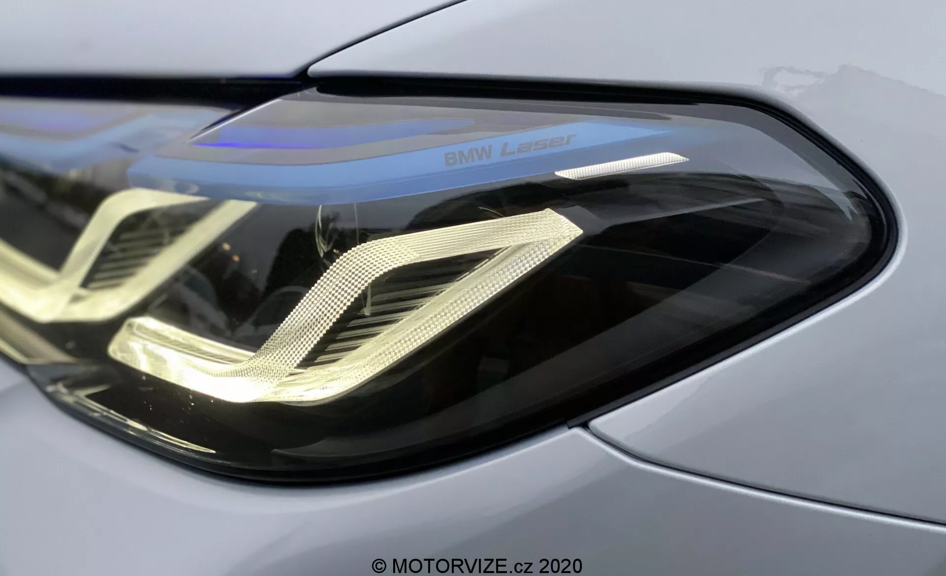 Detailný záber na predný ľavý reflektor BMW 520i Touring Steptronic 2020, s elegantným zakriveným polykarbonátovým krytom šošovky, charakteristickými dennými LED svetlami v tvare dvojitej špirály, odrazovým krytom a označením 'BMW Laser', zdôrazňujúcimi pokročilú laserovú svetelnú technológiu a prémiový kovový panel karosérie.