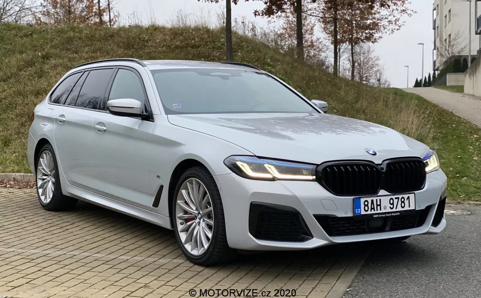 Moderné, luxusné BMW 5 Series (BMW 520i Touring Steptronic) kombi fotografované z nízkeho uhlu trojštvrťového čela. Obrázok zvýrazňuje prednú mriežku automobilu, elegantné a uhlové svetlomety pravdepodobne s LED technológiou a svalnaté dizajnové línie na kapote a blatníkoch. Viditeľné časti zahŕňajú prednú mriežku, nárazník, svetlomety, predné dvere vodiča, predný blatník, časť zadných dverí, bočné okná, kolesá a bočné zrkadlo na strane vodiča. Veľké kolesá z ľahkých zliatin naznačujú športovejšiu verziu modelu a strieborný náter je reflexný a čistý. Auto má výraznú prednú mriežku s vertikálnymi lamelami, odznak na prednom blatníku naznačujúci vyššiu úroveň výbavy a bočné zrkadlá s integrovanými smerovými svetlami. Viditeľný je tiež strešné okno. Vozidlo je zaparkované na mestskej ceste s obrubníkom a chodníkom v popredí a trávnatou oblasťou a budovou na pozadí, pod zatienenými svetelnými podmienkami.