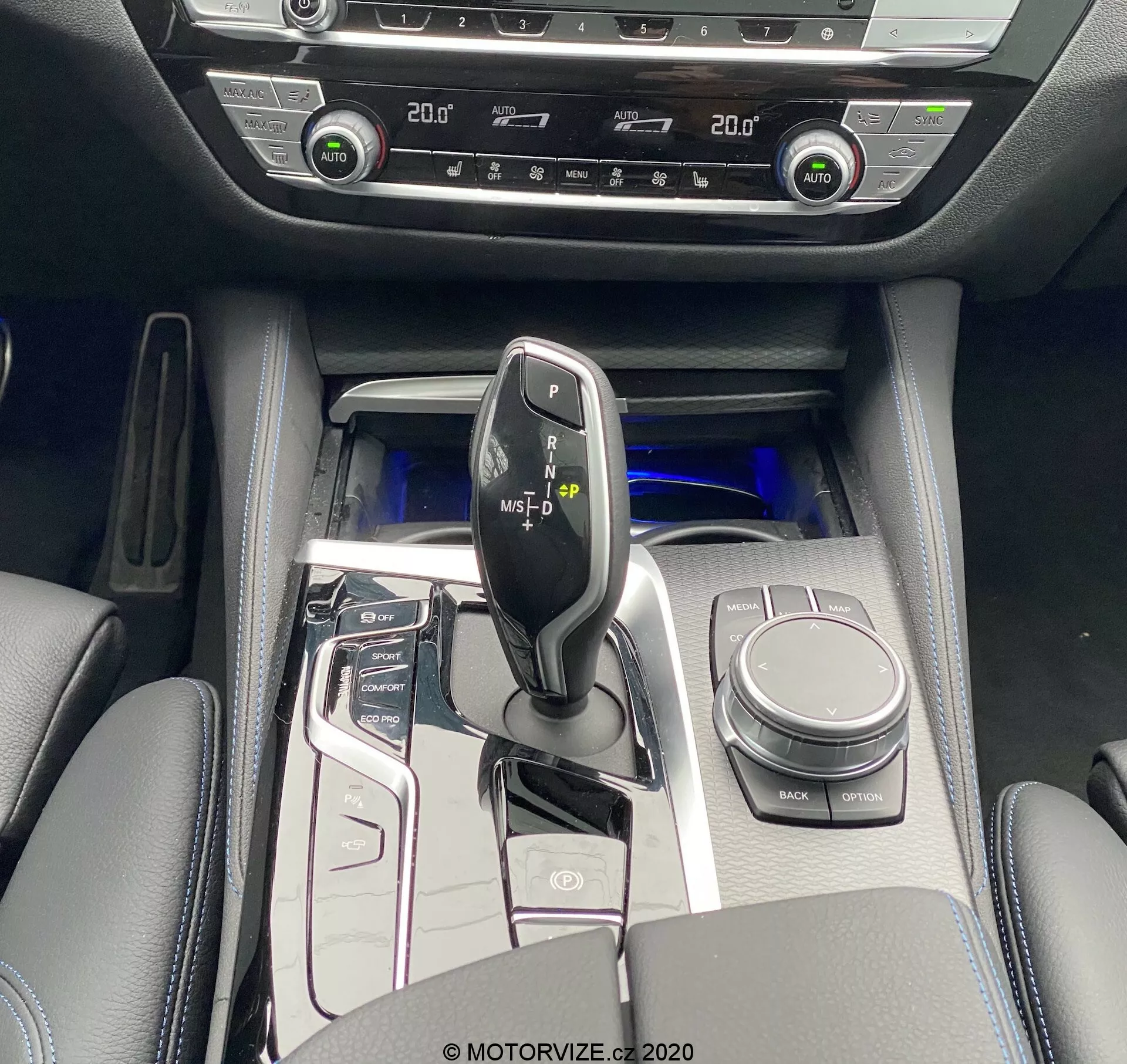 Pohľad z vtáčej perspektívy na centrálnu konzolu BMW 520i Touring Steptronic 2020, zachytávajúci lesklú čiernu radiacu páku s kovovými akcentmi, obklopenú vysoko lesklými ovládacími tlačidlami pre start/stop systém, jazdné režimy, parkovacie senzory a ďalšie. Viditeľný je ovládač iDrive, držiaky na poháre a polstrovaná lakťová opierka s úložným priestorom. Chladivé modré ambientné osvetlenie zvyšuje pocit luxusu. Dvojzónová klimatizácia a ovládanie vyhrievaných sedadiel naznačujú pokročilé komfortné funkcie. Interiérové materiály zahŕňajú povrchy podobné koži s kovovými akcentmi a vysoko lesklými panelmi.