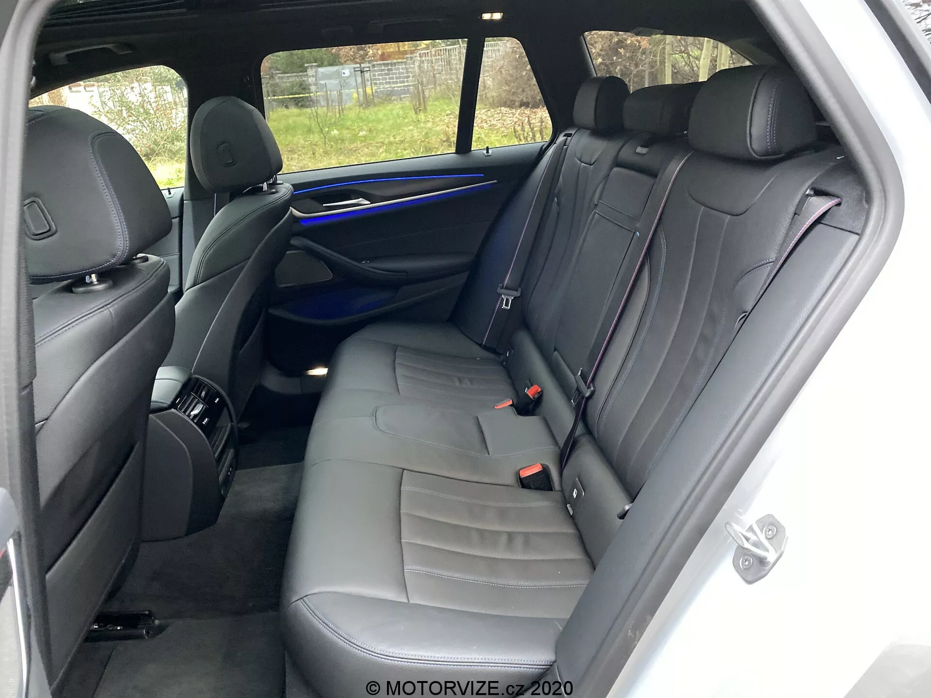 Interiér BMW 520i Touring Steptronic z roku 2020 so zameraním na oblasť zadných sedadiel z pravej strany. Uhol pohľadu je blízko pravých zadných dverí, ukazuje sedadlo pre troch s tmavým koženým čalúnením a dekoračným prešívaním. Funkcie zahŕňajú tri opierky hlavy, tri bezpečnostné pásy, panel dverí s mriežkou reproduktora, rukoväť a ovládacie prvky, modrú ambientnú svetelnú lištu a úložnú sieť na zadnej strane sedadla spolujazdca. Dobre udržiavaný interiér nemá viditeľné osobné veci ani významné opotrebenie. Čiastočný výhľad von cez zadné okno zahŕňa plot.