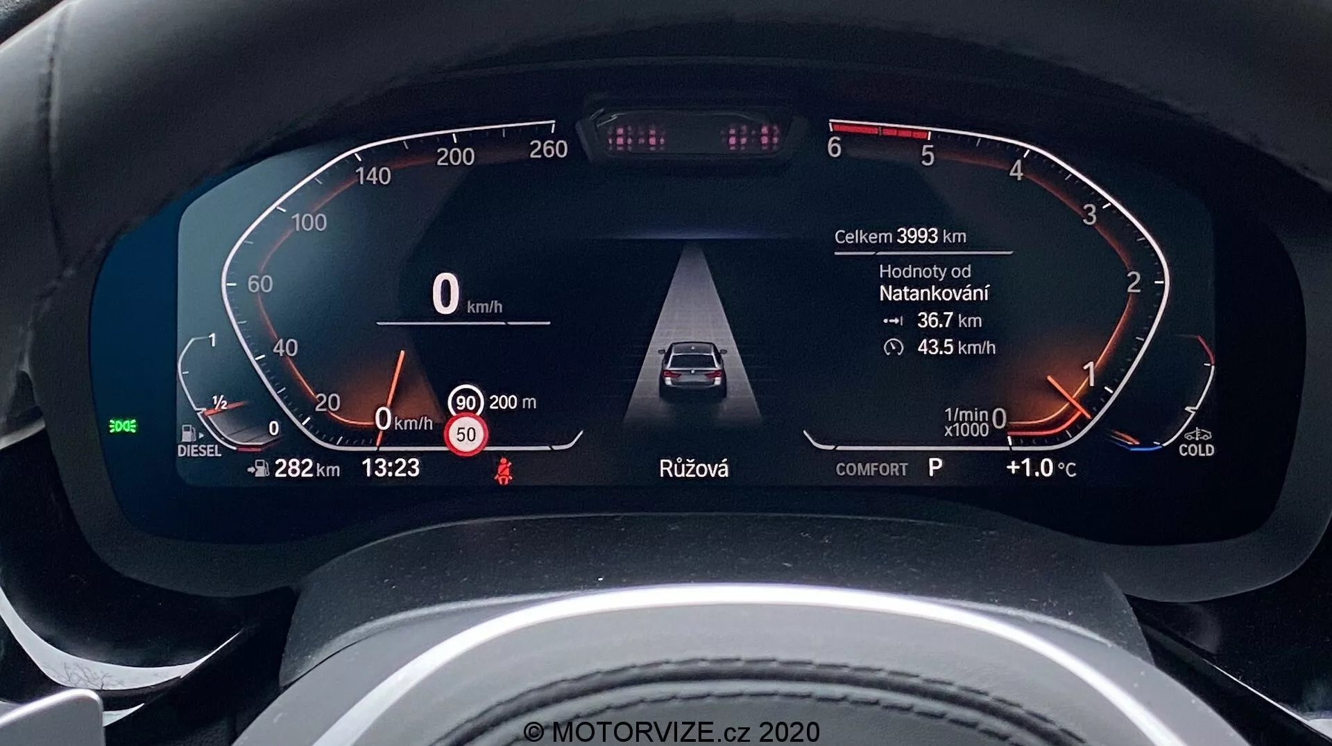 Zblízka na digitálny prístrojový panel BMW radu 5 2020 (BMW 520i Touring Steptronic) z pohľadu vodiča. Digitálny prístrojový displej zobrazuje kľúčové informácie ako rýchlosť vozidla (0 km/h), palivomer označený pre naftu, varovania o otvorených dverách a pripomienky bezpečnostného pásu, pokročilé asistenčné systémy pre vodičov, ako je adaptívny tempomat, otáčkomer v stave voľnobehu s indikátorom studeného motora, a ďalšie údaje, ako napríklad najazdené kilometre (39933 km), vonkajšia teplota (+1.0°C), čas (13:23), výber prevodového stupňa (P pre parkovanie) a režim jazdy (komfort). Dizajn je elegantný a moderný, čo naznačuje luxusné vozidlo.