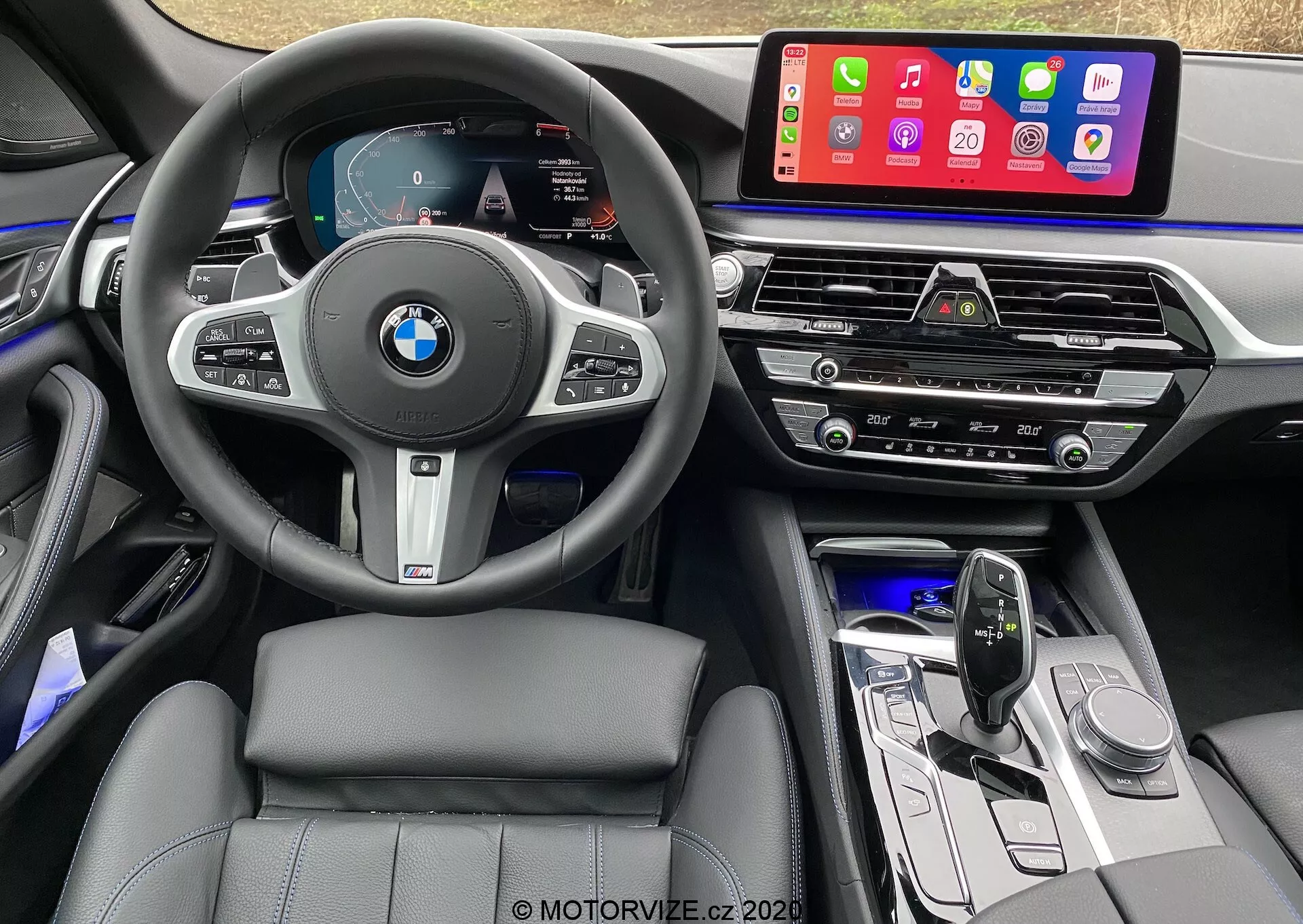 Pohľad do interiéru z miesta vodiča vozidla BMW 520i Touring Steptronic z roku 2020, s koženým multifunkčným volantom s logom BMW a tlačidlami na ovládanie tempomatu, audia a telefonovania. Digitálny prístrojový panel zobrazuje tachometer, otáčkomer a navigačnú mapu. Stredová konzola obsahuje veľký dotykový infotainment systém, ktorý zobrazuje mobilné aplikácie ako Apple CarPlay. Nižšie sú ventilačné otvory, panel klimatizácie, ovládanie audia, moderná radiaca páka a ovládacie tlačidlá. Interiér ukazuje sedadlá s koženým prešívaním, ambientné osvetlenie a vysoko kvalitné materiály s kovovými akcentmi.