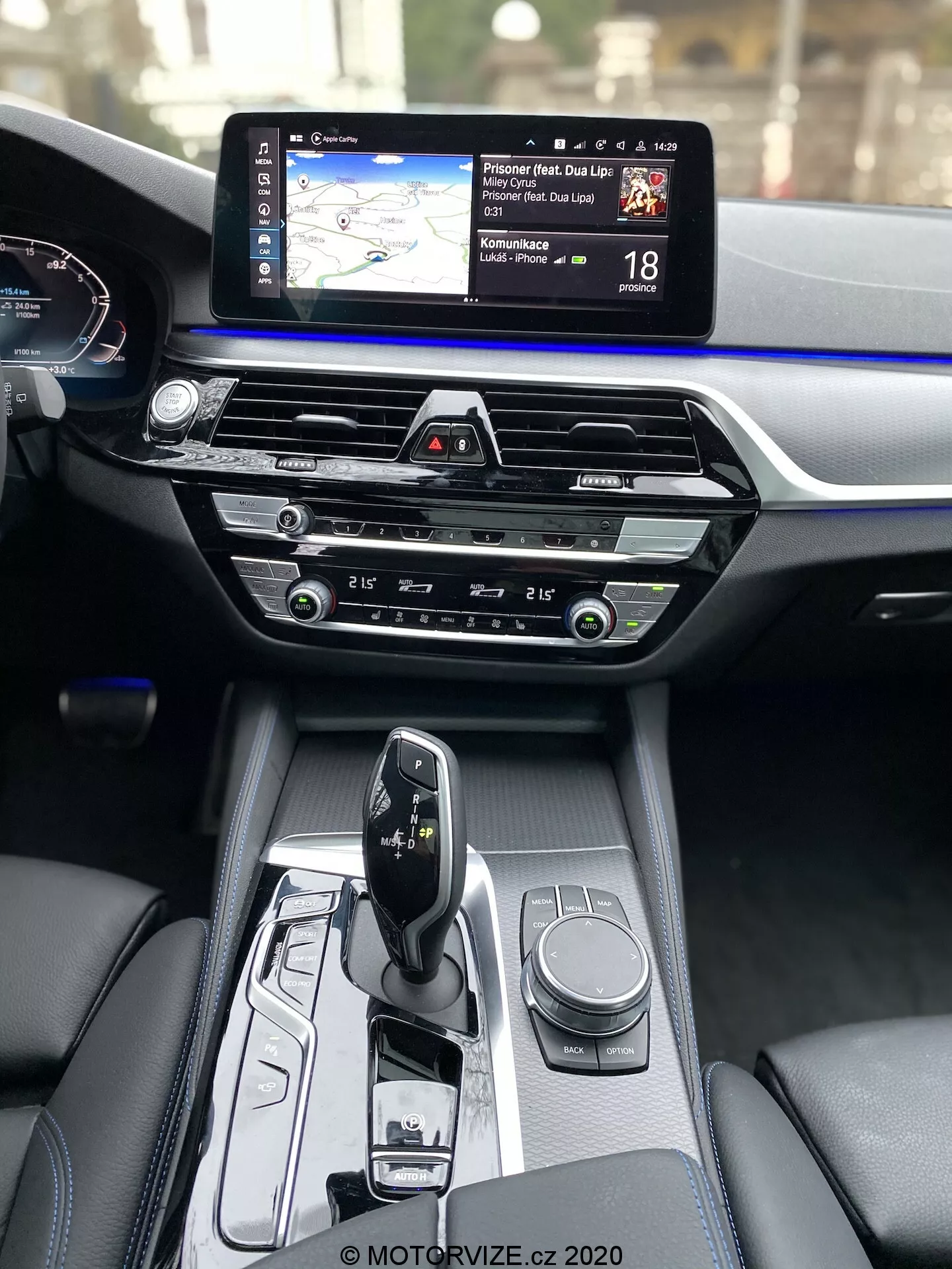 Vnútorný pohľad na BMW radu 5 2020 (BMW 520i Touring Steptronic), zameraný na stredovú konzolu a palubnú dosku z perspektívy predného spolujazdca. Stredová konzola obsahuje moderný volič automatickej prevodovky v režime 'park', s rôznymi ovládacími prvkami vrátane otočného ovládača pre infotainment systém a tlačidiel na jazdné režimy alebo nastavenia vozidla. Stredná časť palubnej dosky zahŕňa ovládacie prvky klimatizácie s intuitívnymi gombíkmi a tlačidlami, nad ktorými je slot pre CD. V hornej časti palubnej dosky je výrazne viditeľný široký dotykový displej infotainmentu s navigačnou mapou a informáciami o rýchlostných obmedzeniach. Čiastočne viditeľný je digitálny prístrojový panel zobrazujúci rýchlomer. Materiály interiéru zdôrazňujú prémiové spracovanie, kontrast medzi tmavou kožou a modrým ozdobným lemovaním naprieč celou šírkou palubnej dosky.