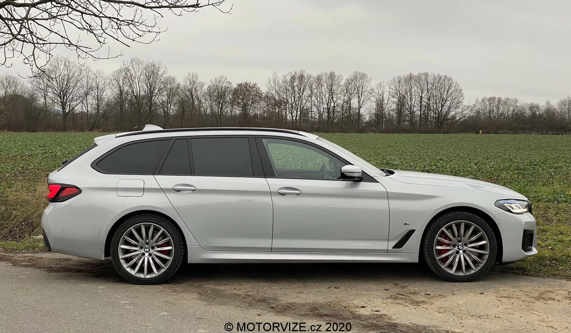 Bočný profil moderného BMW 520i Touring Steptronic kombi zachyteného počas denného svetla, ukazujúci elegantné dizajnové línie, päť dverí vrátane zadných, dlhú strechu so strešným spojlerom, veľké viaclúčové zliatinové kolesá s viditeľnými brzdovými strmeňmi, kľučky dverí vo farbe karosérie, integrované smerovky v spätných zrkadlách, ľahko zatmavené okná a matný alebo nemetalický povrch farby. Auto je umiestnené na štrkovom alebo blatovom ramene vedľa asfaltovej cesty v vidieckom prostredí, s trávou a stromami v pozadí, za priaznivých poveternostných podmienok.
