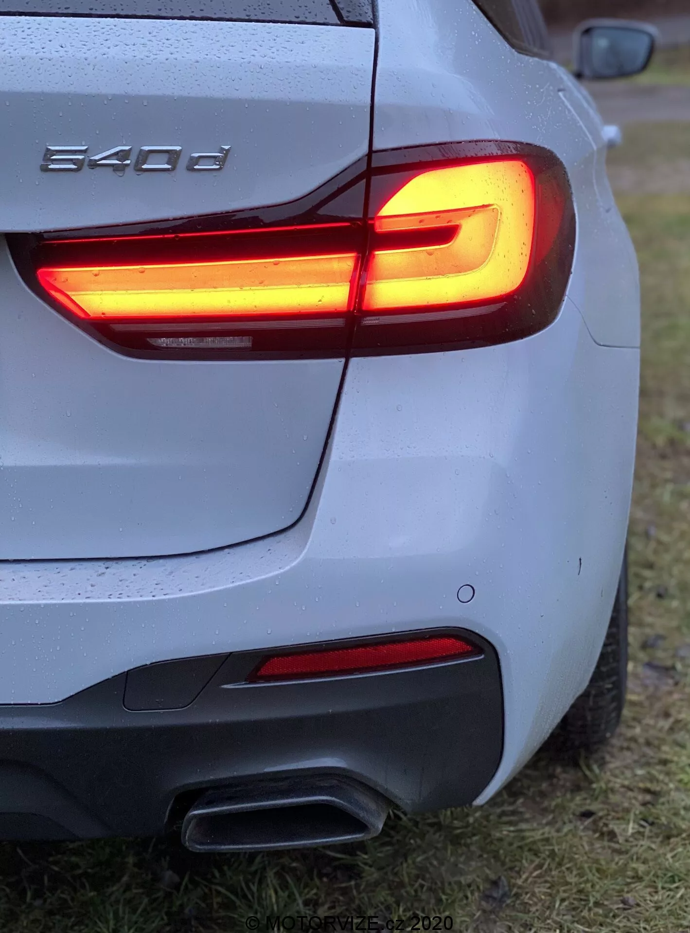 Detailný pohľad na zadný trojštvrtinový uhol BMW 540i, ukazujúc LED zadné svetlo v tvare L, dvojité výfukové rúry, strieborné/sivé telo s kvapkami vody, tvarovaný zadný nárazník, čiastočné logo BMW, zaparkované na trávičnatej ploche.