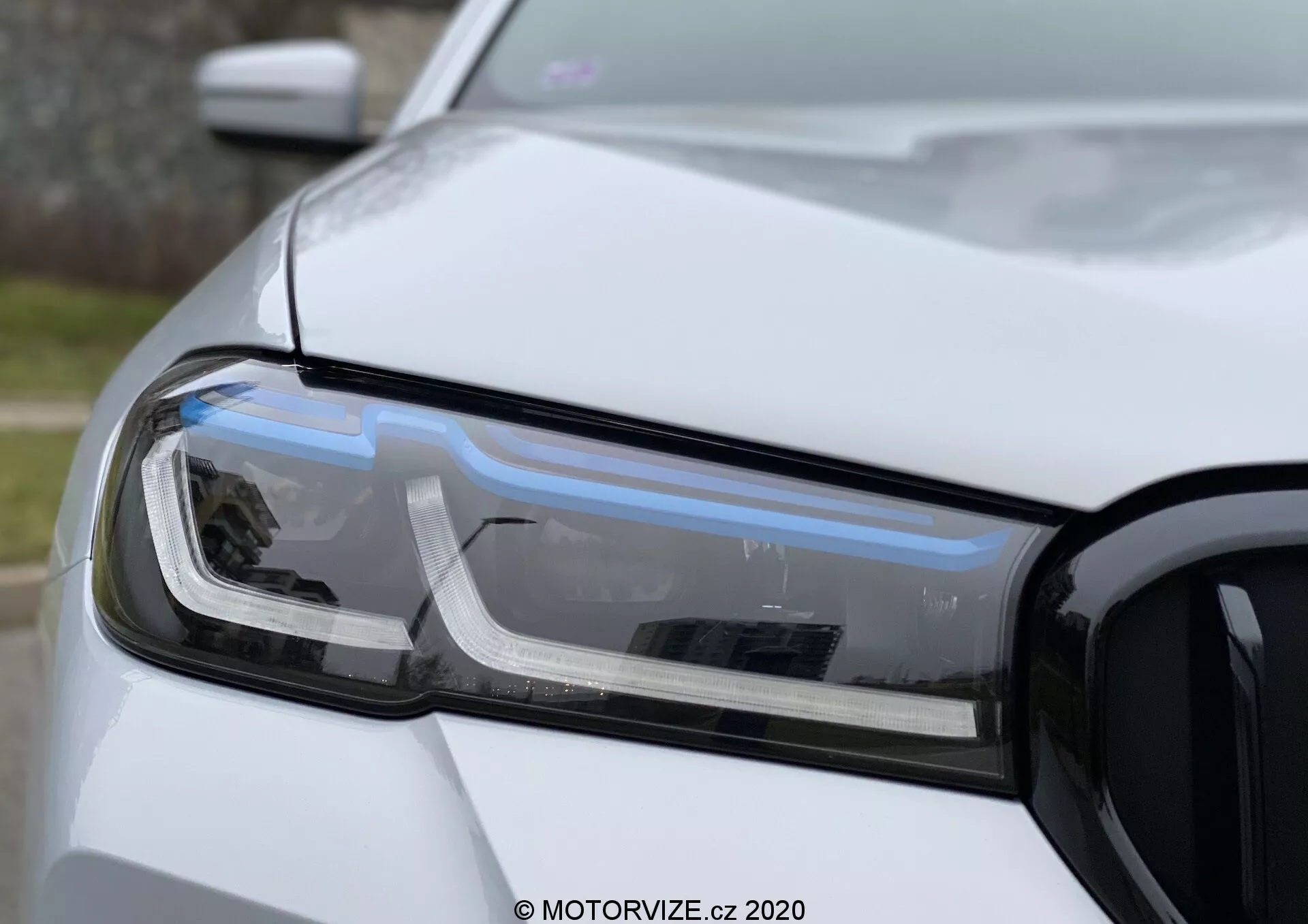 Makro pohľad na svetlomet na strane vodiča BMW 520i Touring Steptronic, so zameraním na LED denné svetlá, projektorový svetlomet a smerovku. Fotografia zobrazuje ľavú prednú časť auta vrátane časti kapoty, predného nárazníka a blatníka, s mriežkou viditeľnou napravo. Záber s nízkou hĺbkou ostrosti vyzdvihuje technológiu a dizajn svetlometu v prírodnom difúznom svetle s pevnou, nemetalickou bielou farbou.