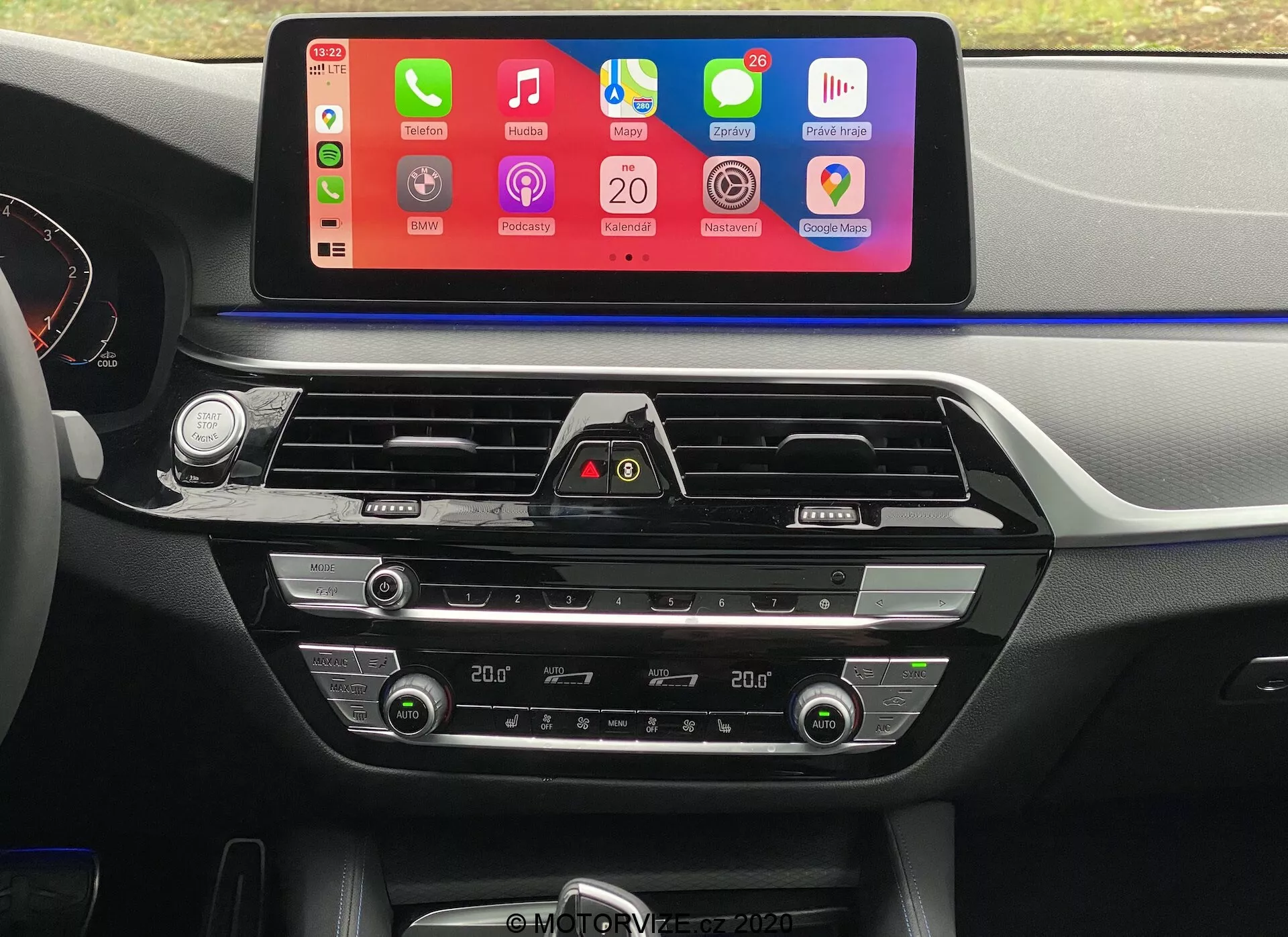 Interiérový záber z BMW 5er-Reihe 2020 (BMW 520i Touring Steptronic) z perspektívy vodičovho sedadla, zameriavajúci sa na centrálny prístrojový panel. Najdominantnejším prvkom je veľký, horizontálne orientovaný dotykový displej infotainmentu s farebným rozhraním podporujúcim integráciu smartfónov ako Apple CarPlay a Android Auto. Pod obrazovkou sú viditeľné vetracie otvory s chrómovými akcentmi a tlačidlo výstražných svetiel. Nižšie sú ovládače klimatizácie s digitálnymi displejmi zobrazujúcimi nastavenie teploty v kabíne, tlačidlá a ovládače na nastavovanie teploty vzduchu, rýchlosti ventilátora a distribúcie vzduchu. Materiály použité na prístrojovom paneli a stredovej konzole obsahujú kombináciu mäkkého plastu a kovových akcentov, čo zvýrazňuje estetický vzhľad a naznačuje dôraz na kvalitu v dizajne interiéru auta.