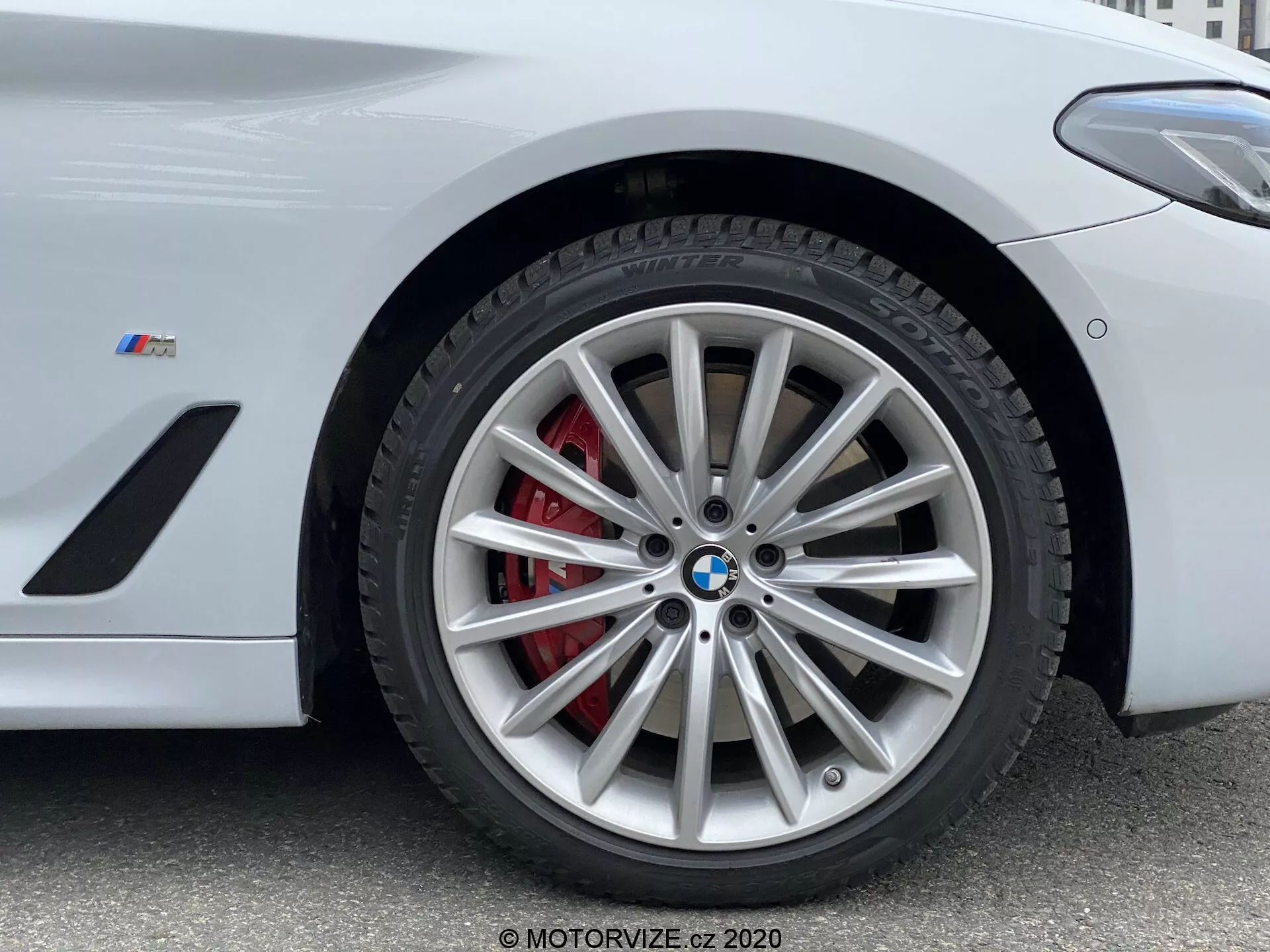 Detailný pohľad na prednú časť kolesa BMW zo série 5 z roku 2020, konkrétne model 520i Touring Steptronic. Fotografia z nízkeho uhla zvýrazňuje zložitý dizajn zliatinového kolesa s mnohými lúčmi a strieborným povrchom, doplnený červenou brzdovou strmeňou a ventilovaným rotorom. Znak BMW a 'M' odznak na blatníku naznačujú, že ide o výkonné vozidlo z BMW M série. Obrázok zachytáva kľúčové detaily vhodné na predstavenie vysokovýkonných brzdových systémov, dizajnu luxusných vozidiel a technológie pneumatík.