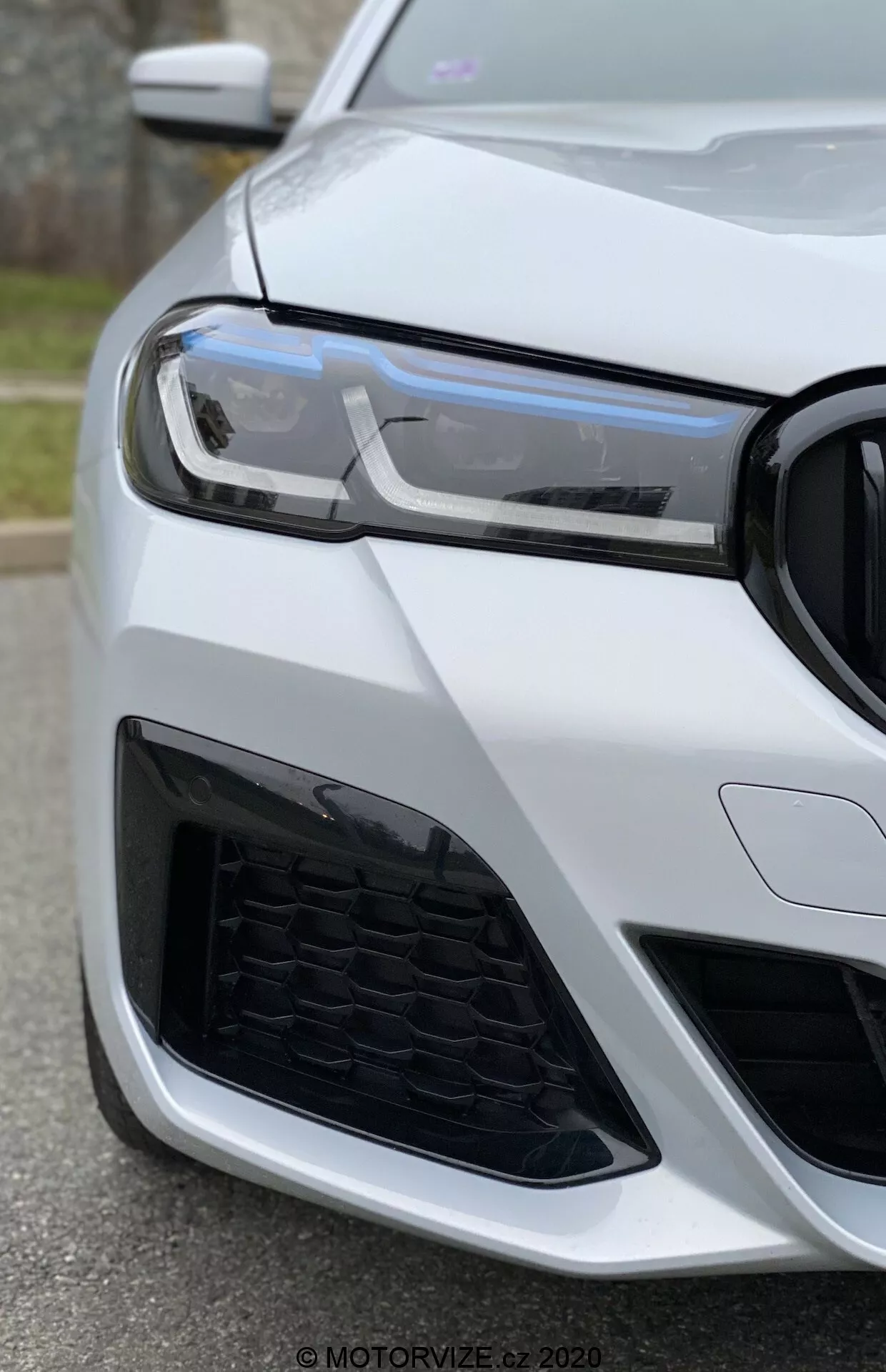 Detailný pohľad na predný ľavý roh BMW 520i Touring Steptronic (2020) z nízkeho uhla, zameriavac sa na svetlomet, mriežku a nárazník. Moderné LED denné svetlá, voštinový vzor mriežky a agresívny dizajn nasávania vzduchu naznačujú športové a pokročilé vozidlo. Auto je natreté lesklou bielou farbou, čo zdôrazňuje jeho luxusný a dobre udržiavaný vzhľad.